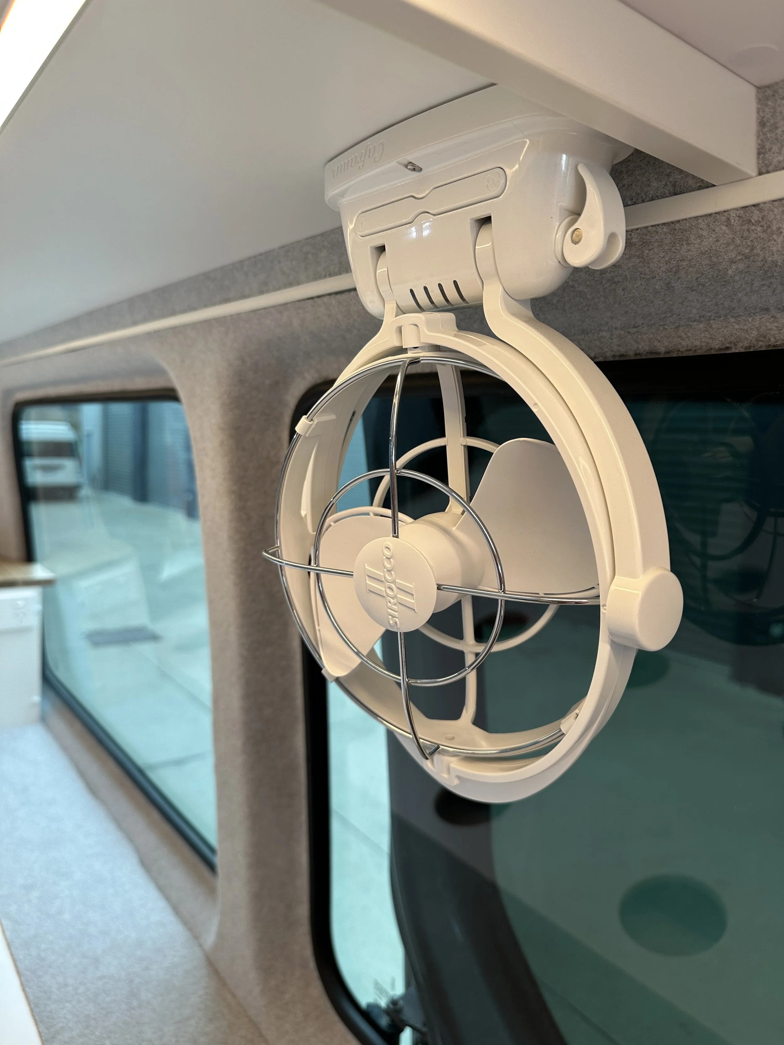 A small white fan with a protective metal grill hanging inside a vehicle near a window.