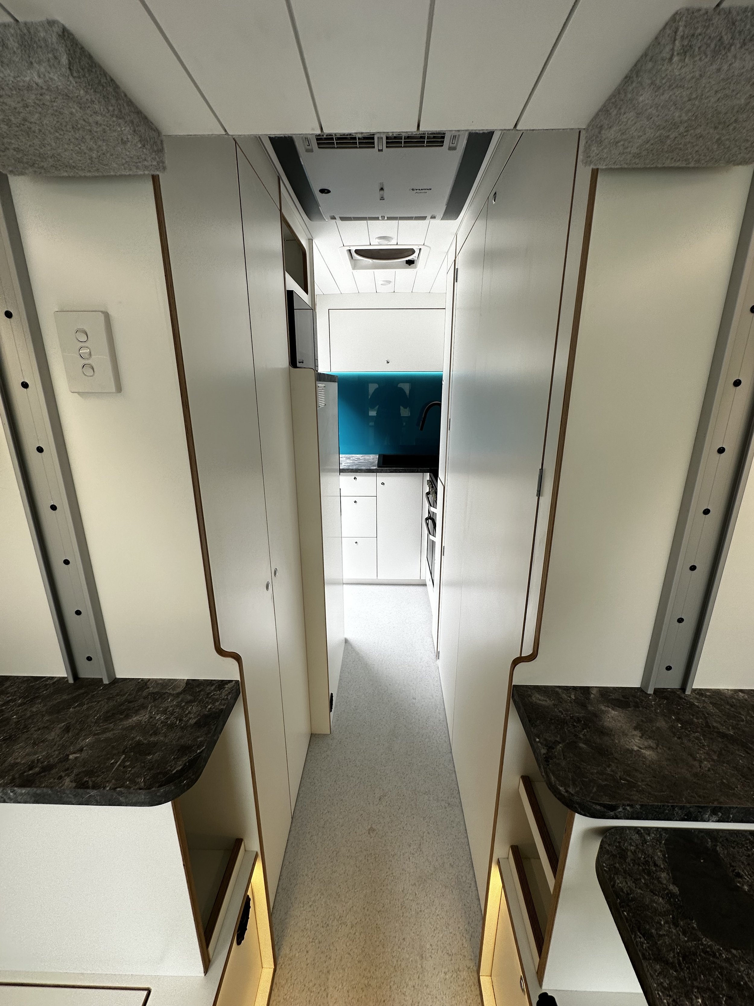 Interior view of a small kitchen area with white cabinets, a black countertop, and a blue backsplash, with a narrow walkway and ceiling vent.