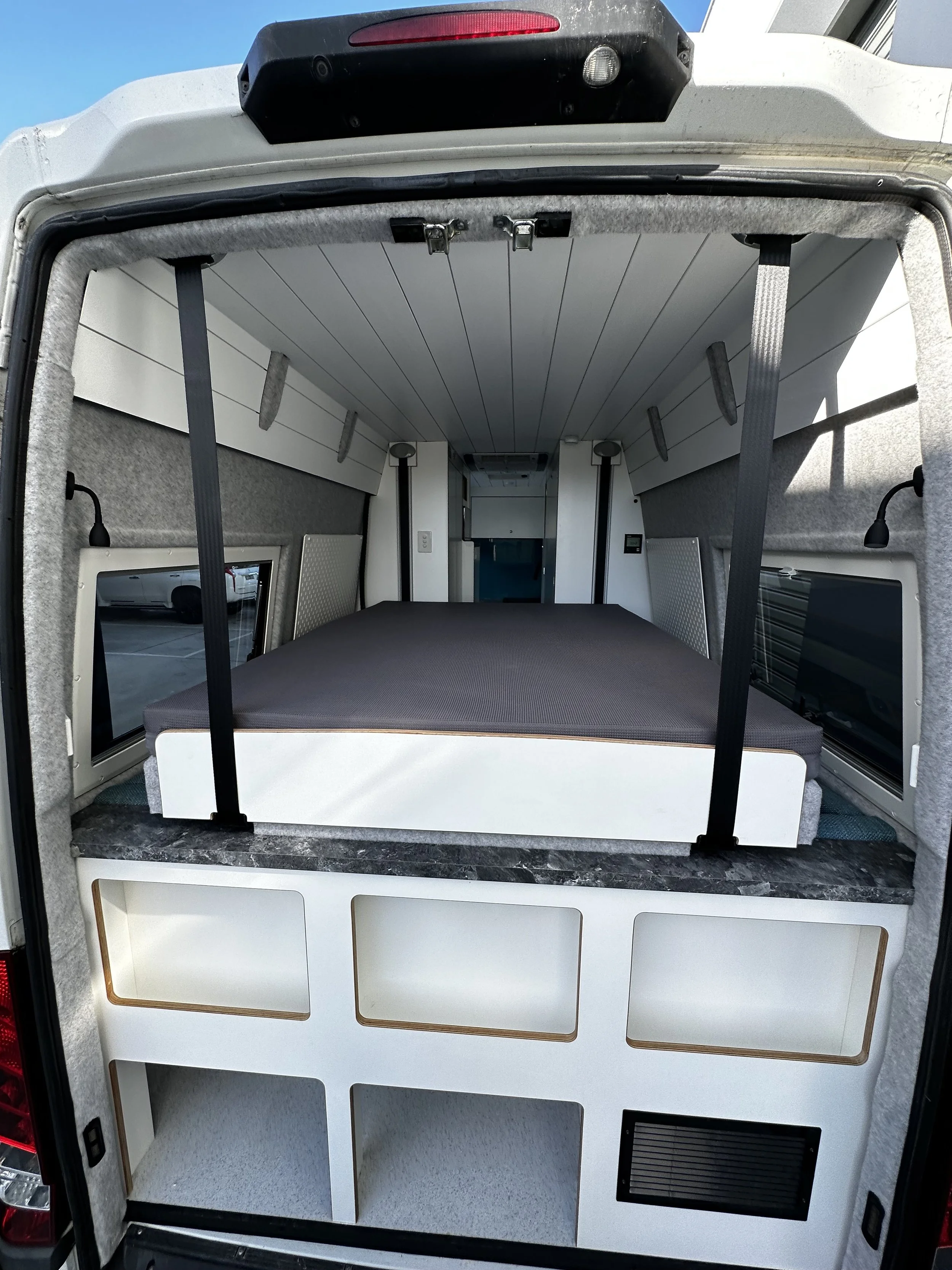 Interior of a camper van with a bed, small windows, and storage compartments.