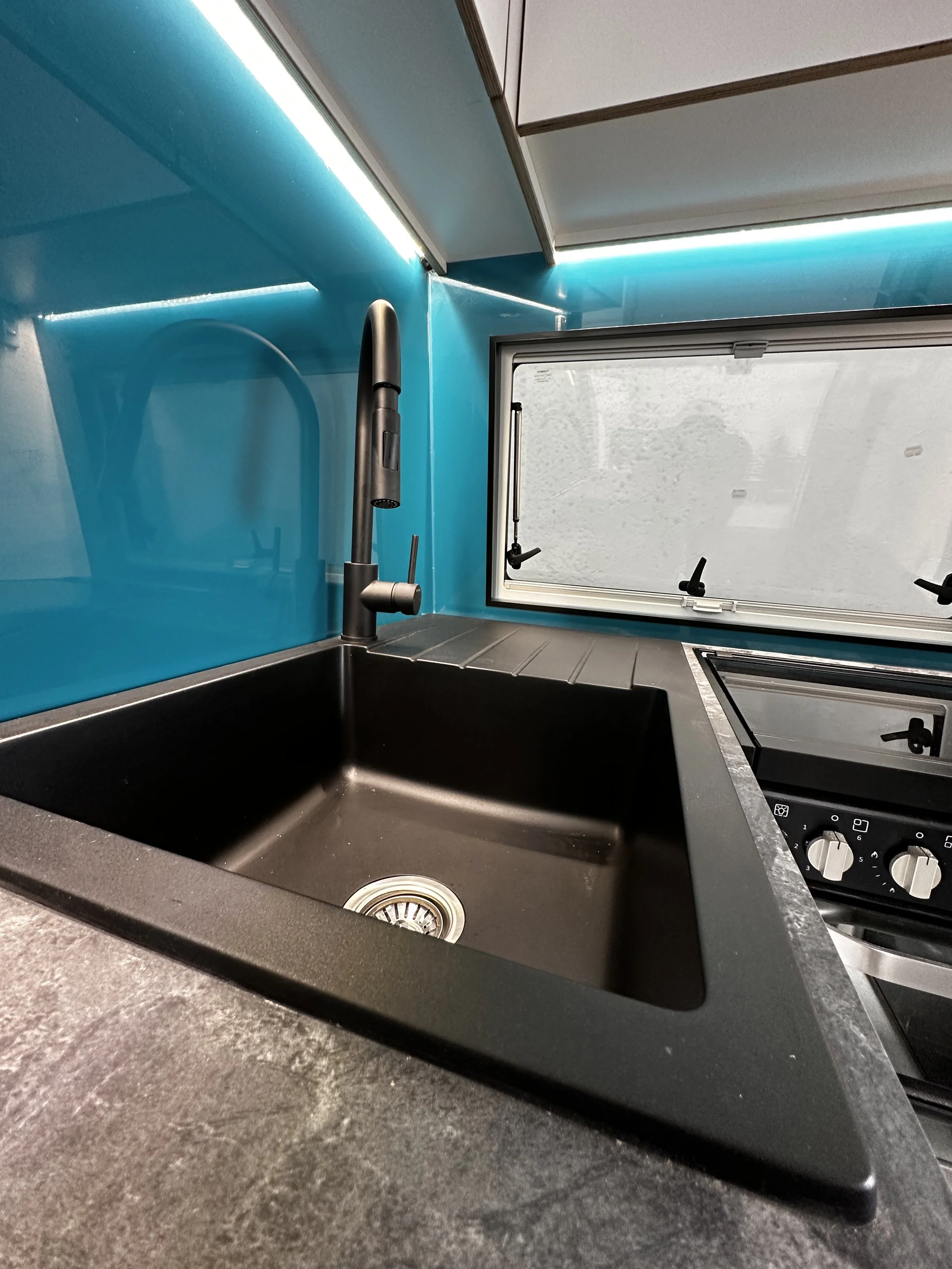 Black kitchen sink with a modern black faucet on a dark countertop, blue wall, small window with handles, and a stove with knobs.