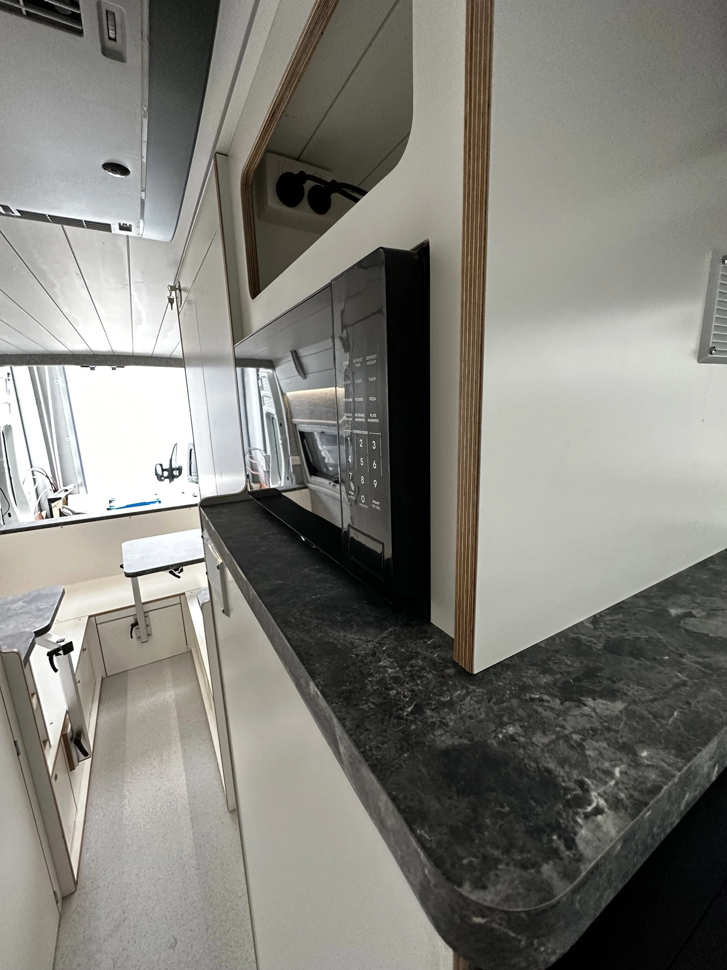 A kitchen with a black microwave oven on a dark granite countertop, light-colored cabinetry, a window with blinds, and kitchen appliances visible through the window.
