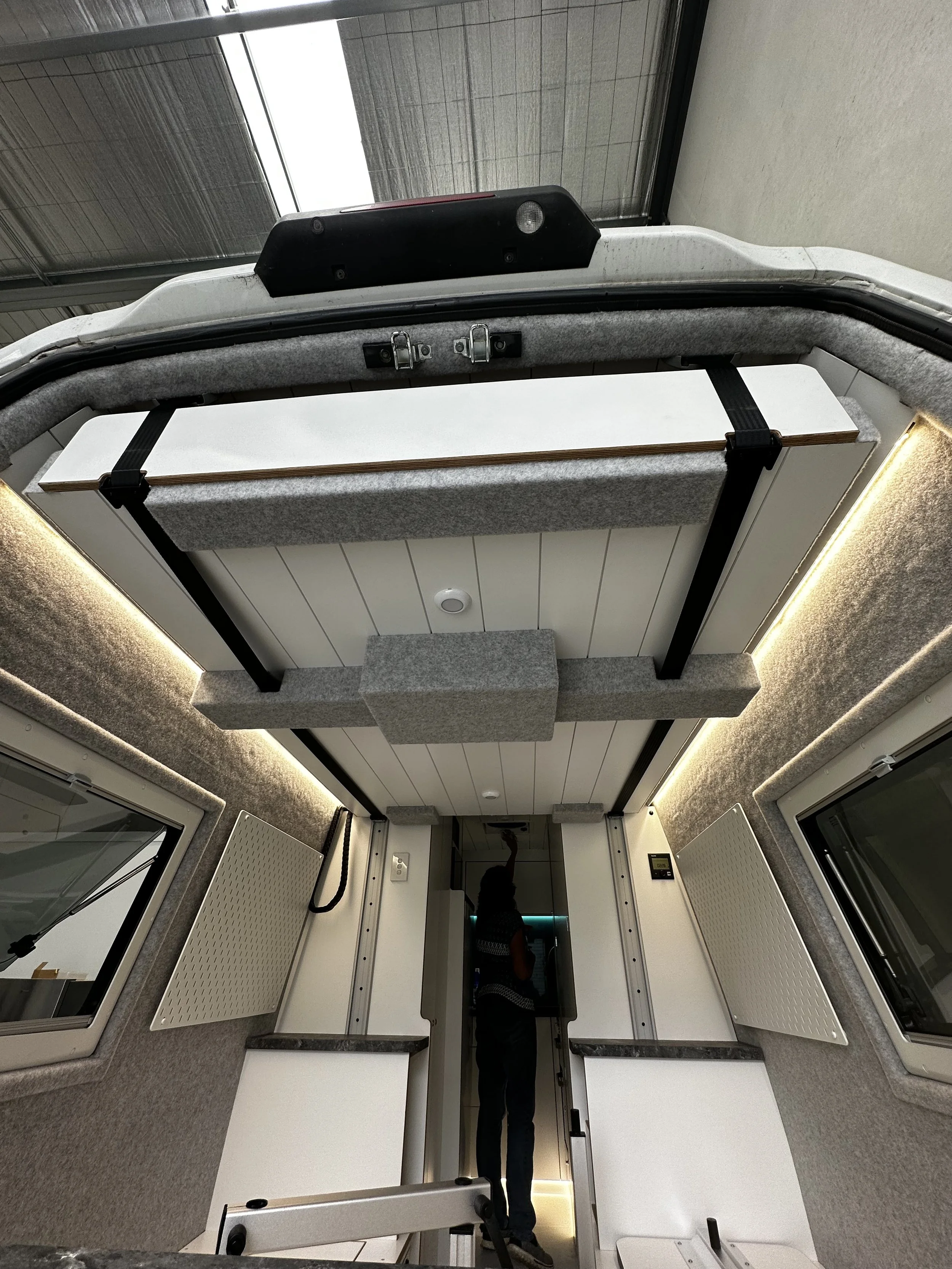 Interior view of a converted van showing ceiling storage compartment, insulated walls, windows, and a person standing by the door.