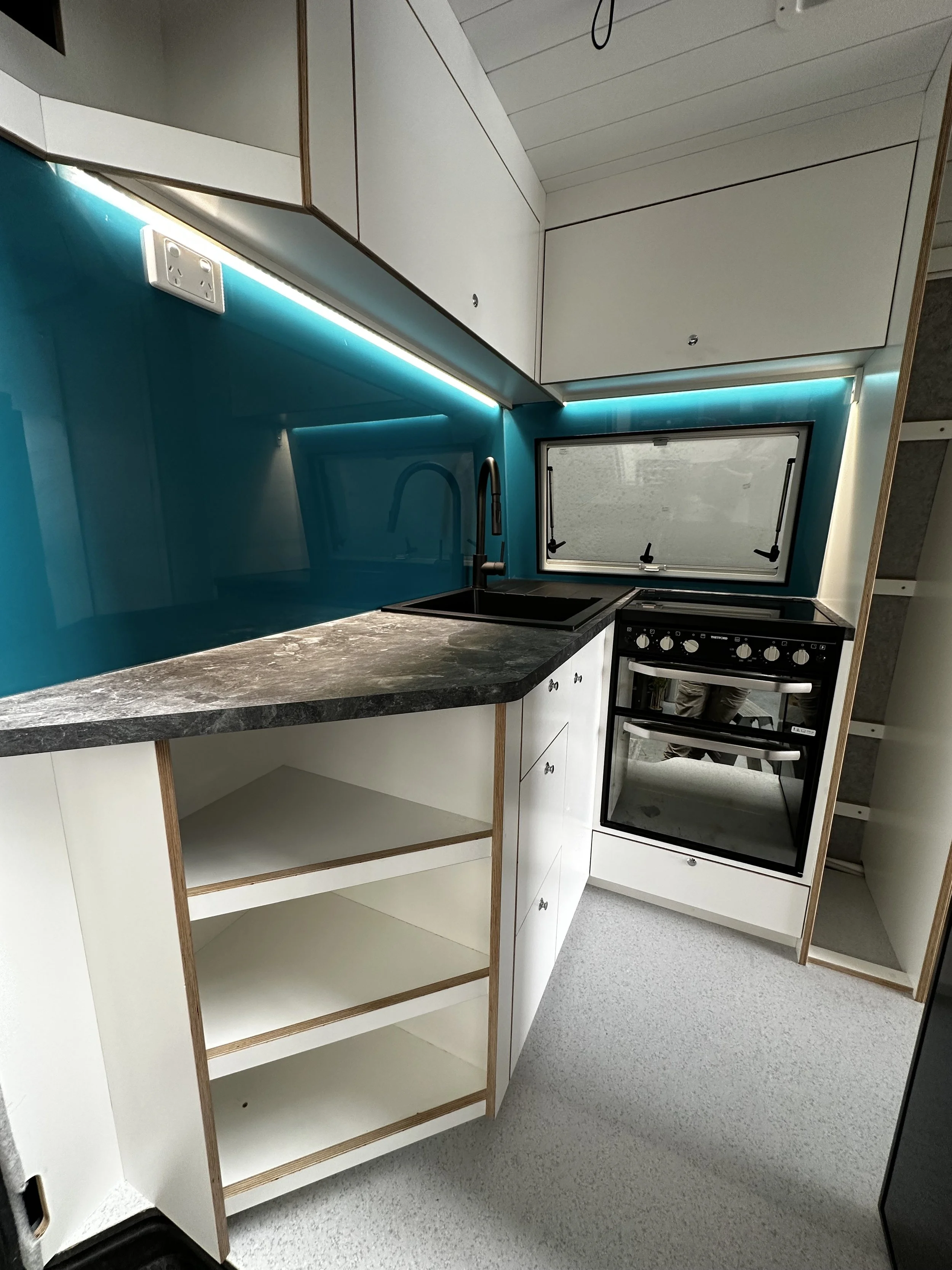 Modern kitchen with white cabinets, black marble countertop, blue glass backsplash, black sink with a tall black faucet, window above the sink, black oven with knobs, open shelving below the countertop, and LED strip lighting under upper cabinets.