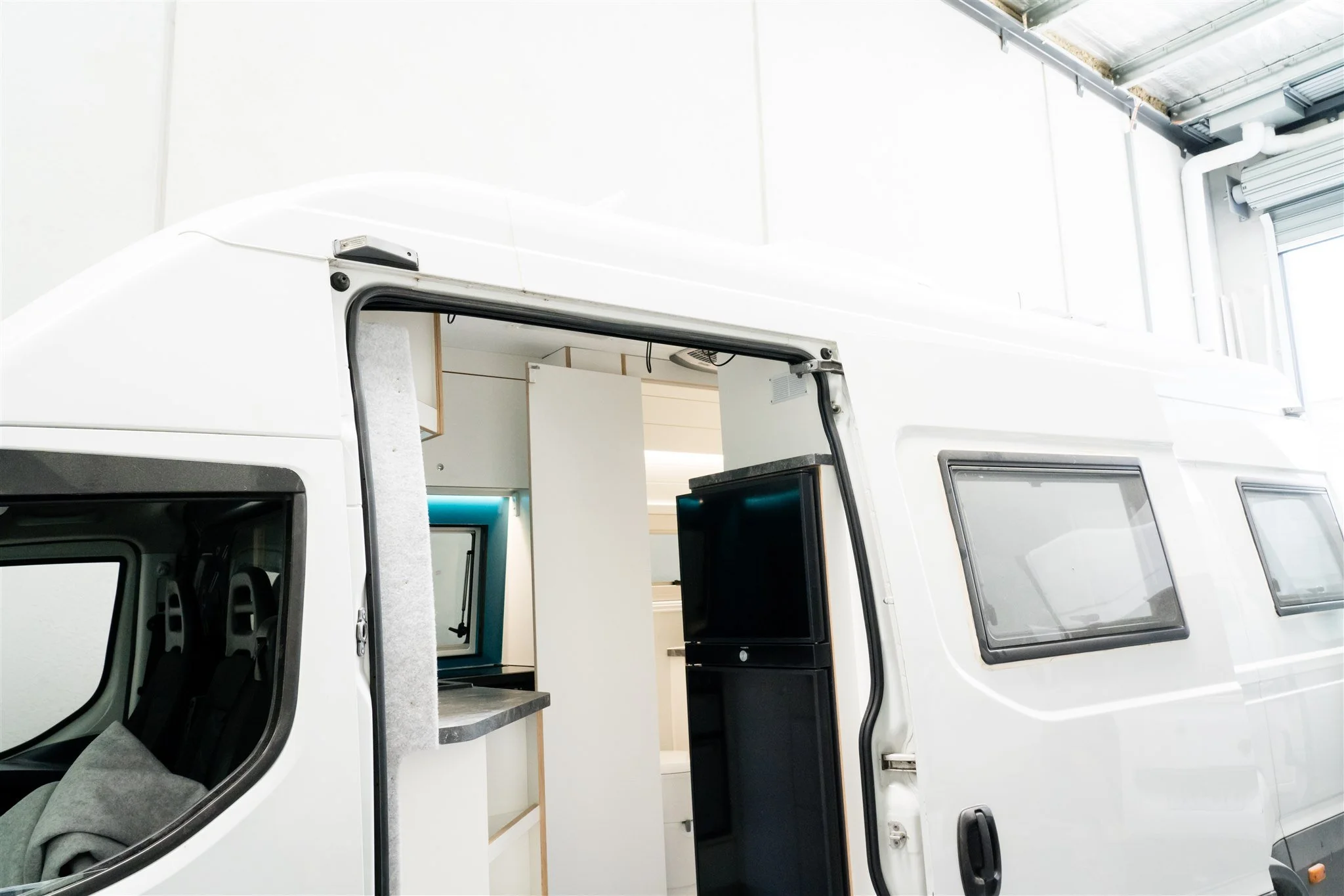 Interior view of a modern white camper van with open side door, showing a small workspace with a desk and window, a refrigerator, and part of a seating area.