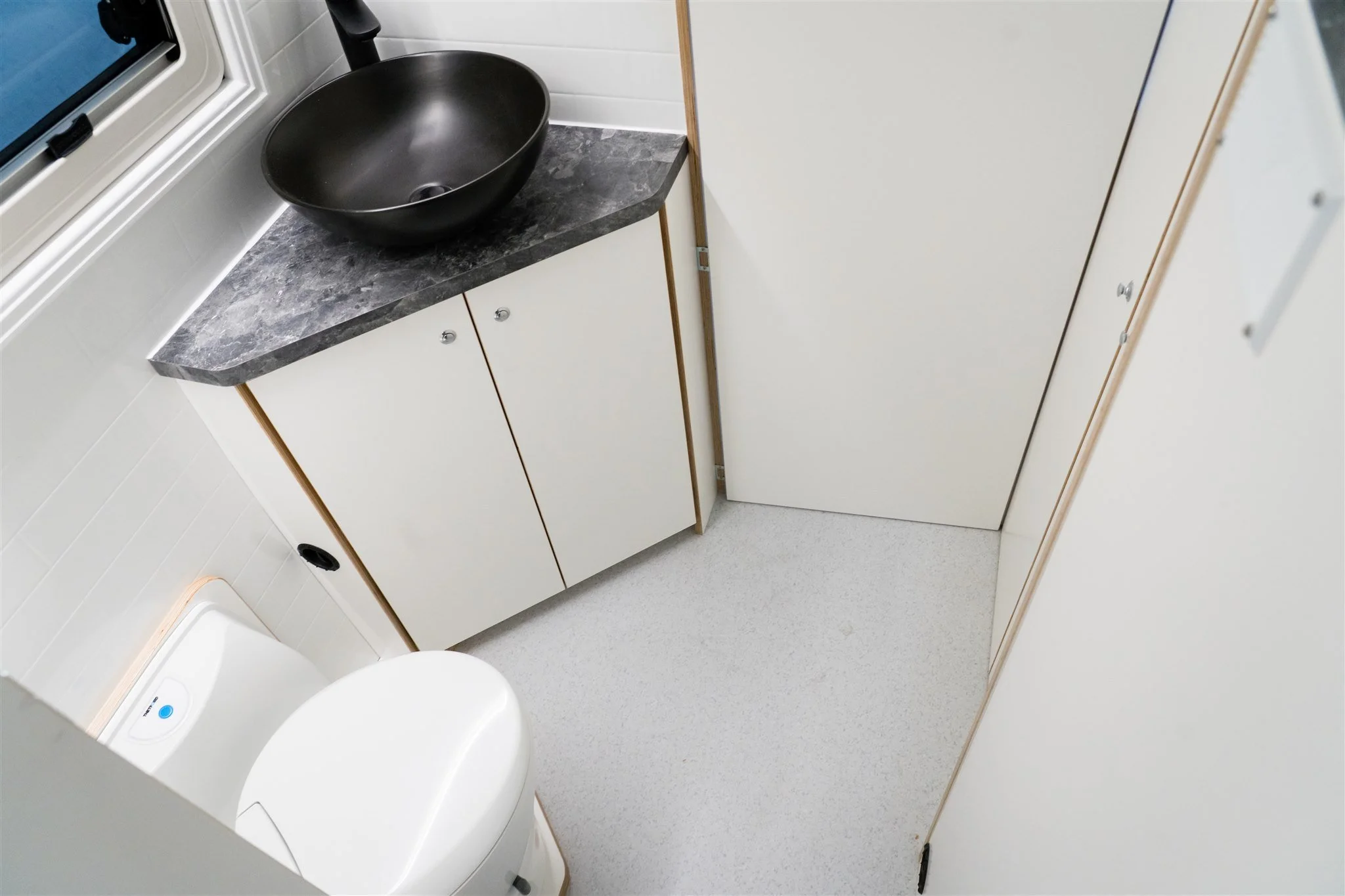 A small bathroom or washroom with a black round sink on a dark gray marble countertop, white cabinet below, a white toilet, and a door. There is a window above the sink.