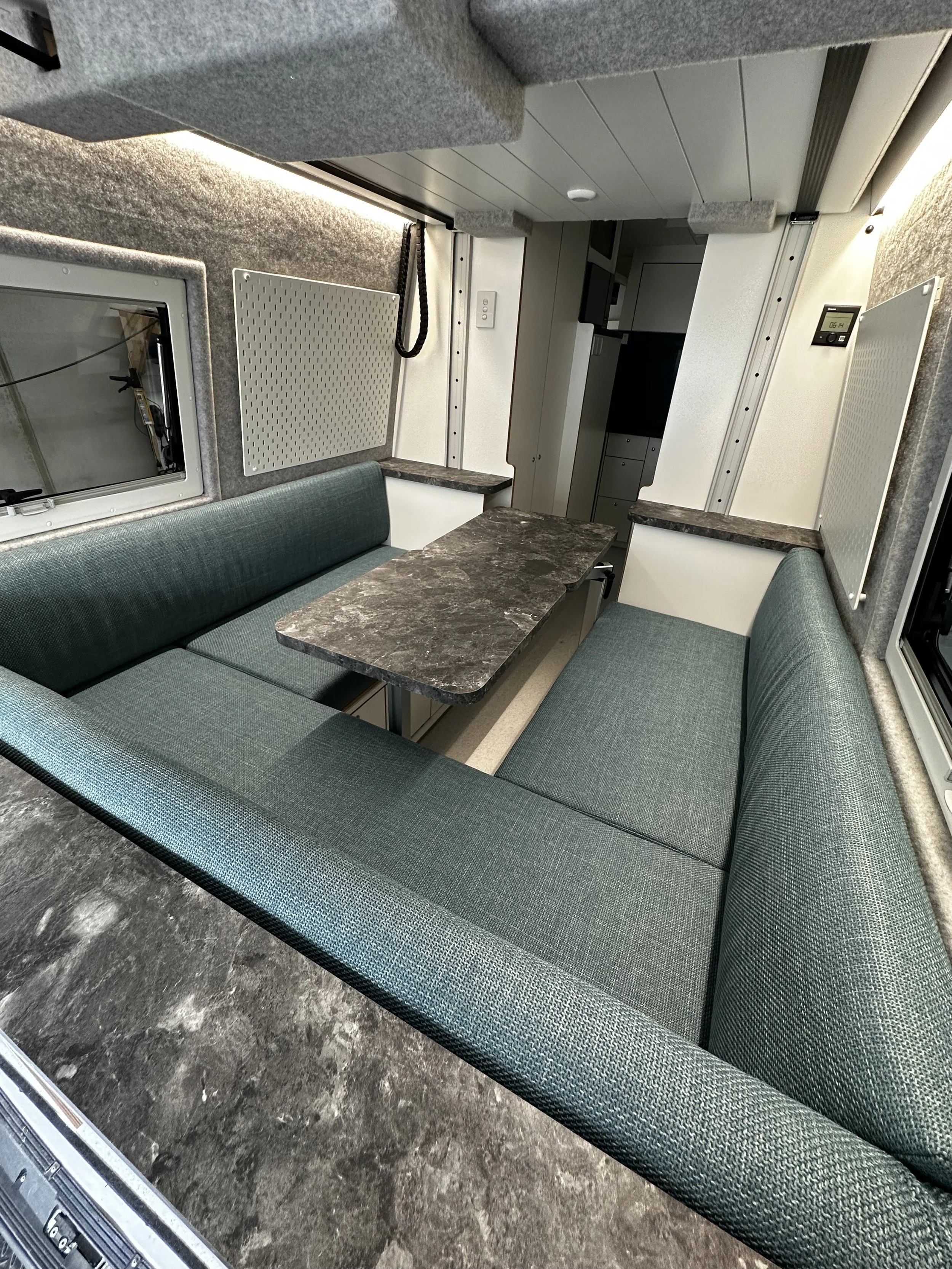 Interior of a small, enclosed space with green cushioned seating arranged in a U-shape around a dark gray marble table, with gray walls, small windows, and various panels and electrical outlets.