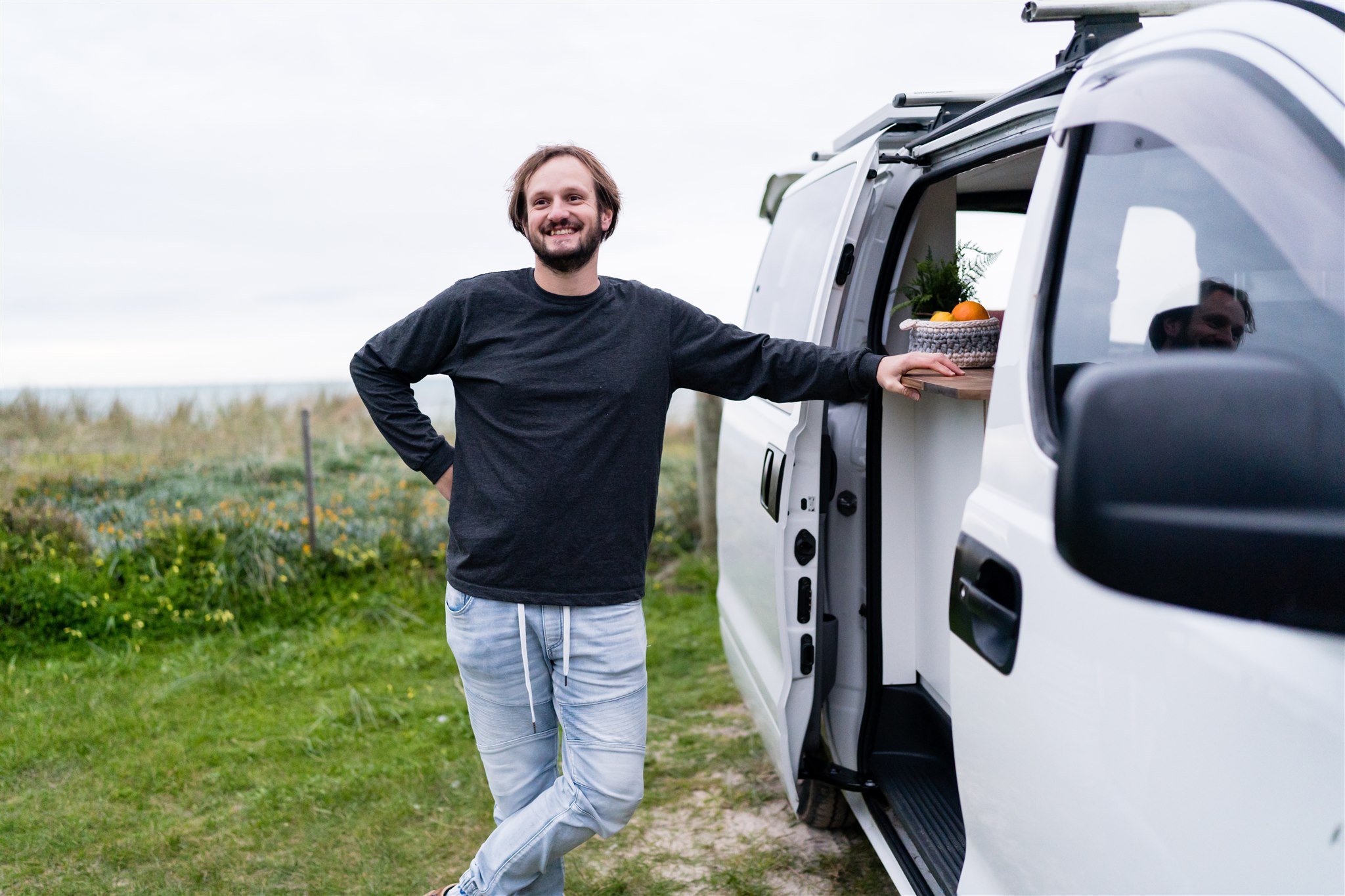 A man standing outside a white van with an open sliding door, smiling and leaning on a wooden counter with a basket of oranges and a potted plant, on a grassy field.