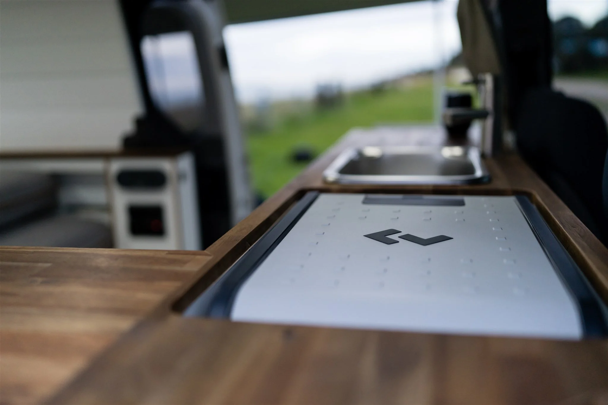 Interior of a camper van with a wooden countertop, a sink, and a digital cooktop, set against a blurred outdoor landscape.