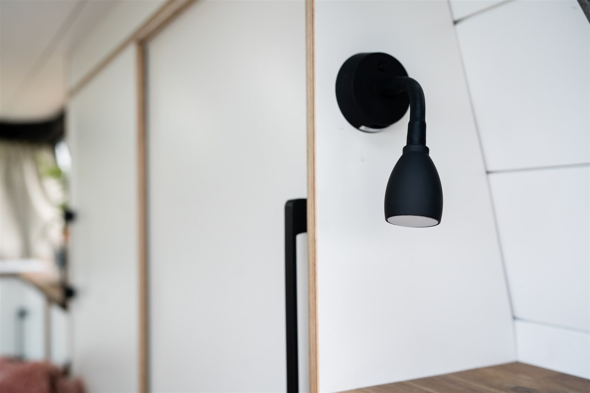 Black wall-mounted light fixture in modern interior, with a white wall and wooden ceiling detail in the background.