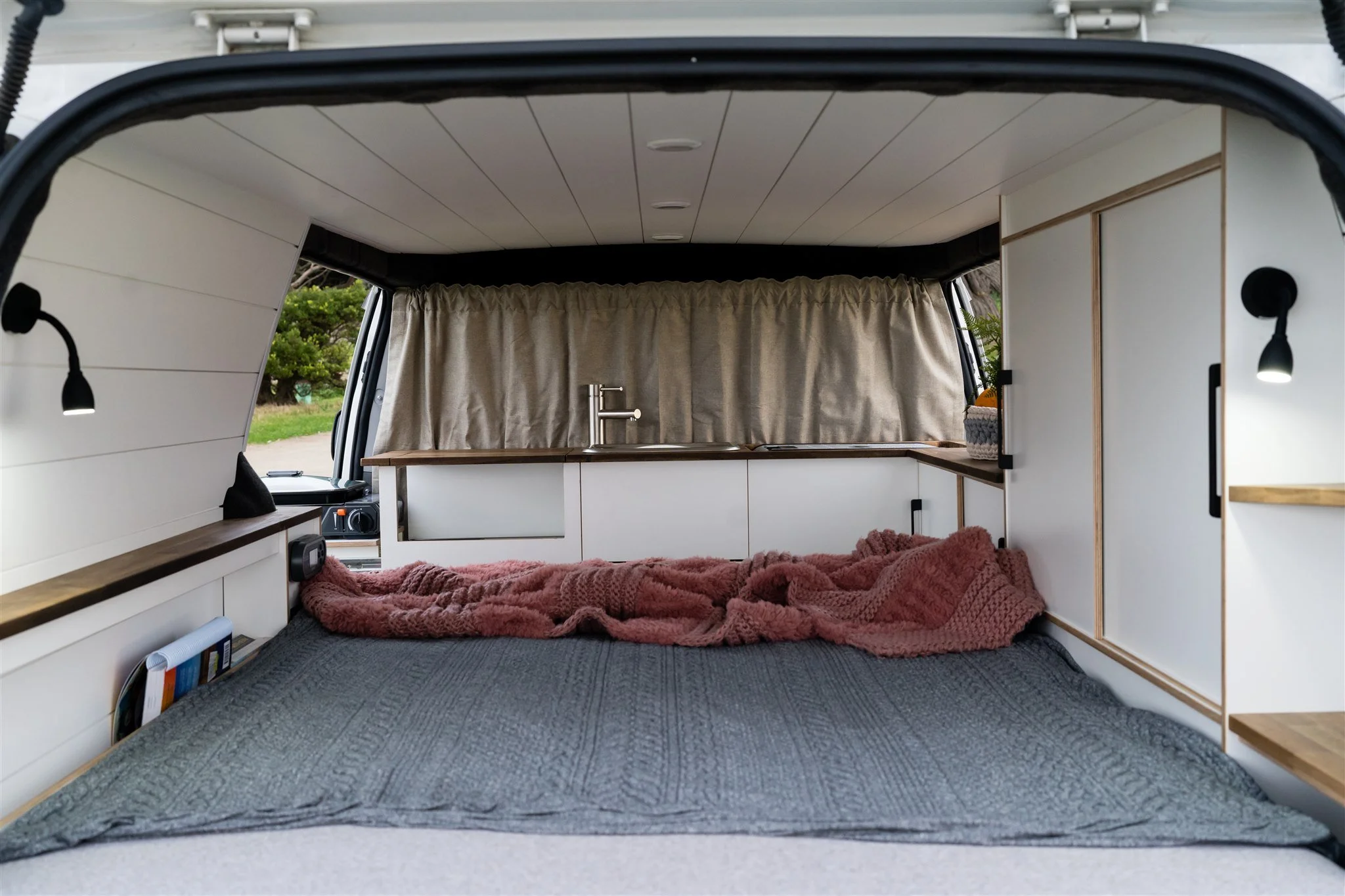 Interior of a camper van with a bed, pink blanket, kitchen sink, beige curtain, and small shelves.