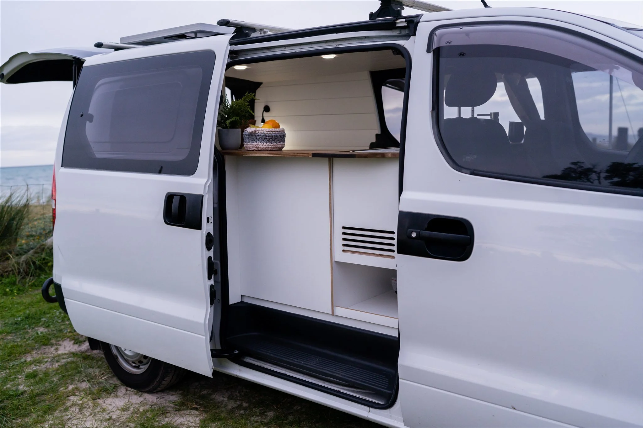 Interior of a white camper van with a small kitchen area, including a wooden countertop, a potted plant, and a bowl of fruit, parked near a beach.