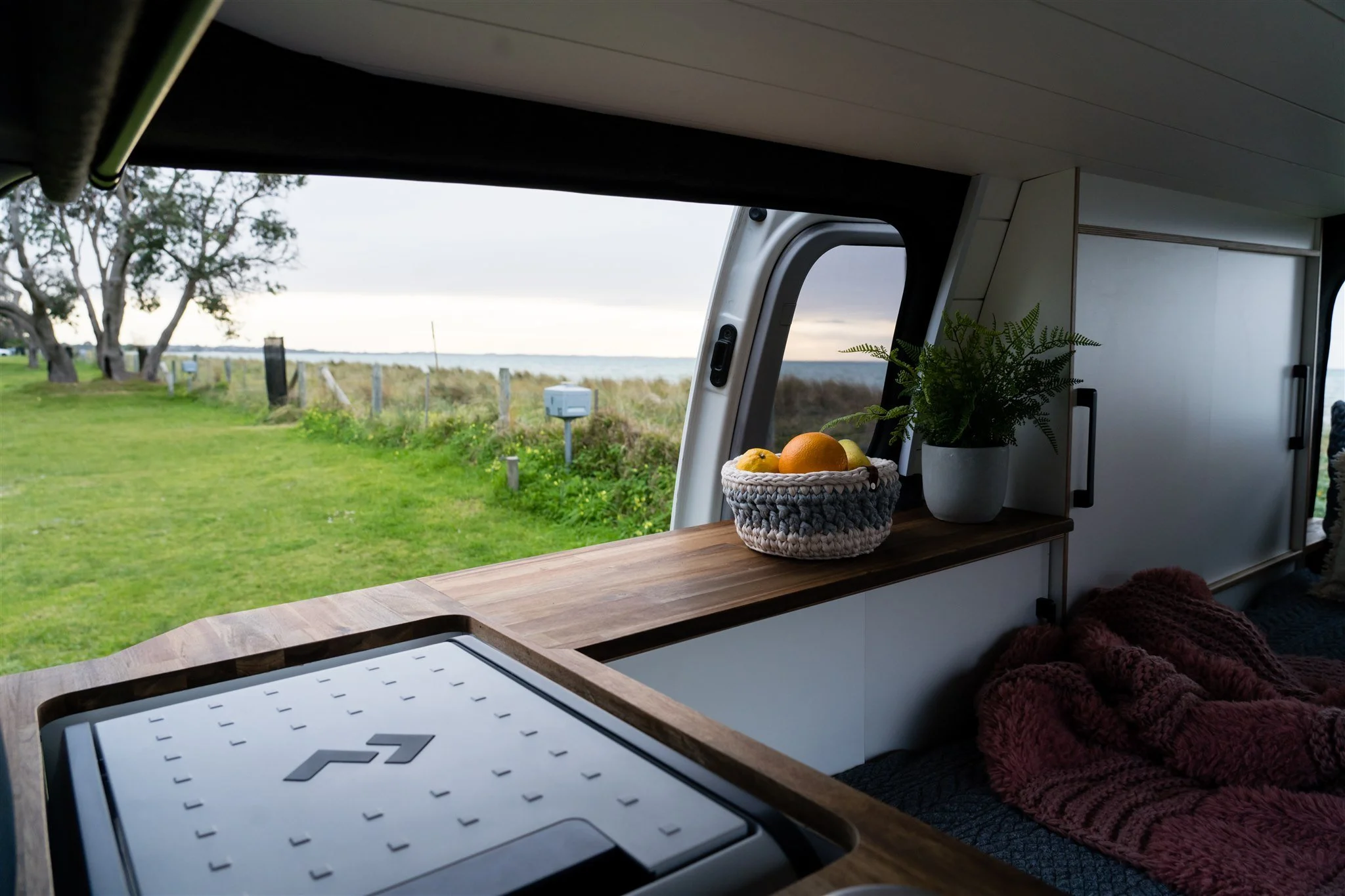 Interior of a camper van with a countertop holding a basket of oranges and a potted plant, looking out onto a grassy landscape with trees, a fence, and a body of water in the background.
