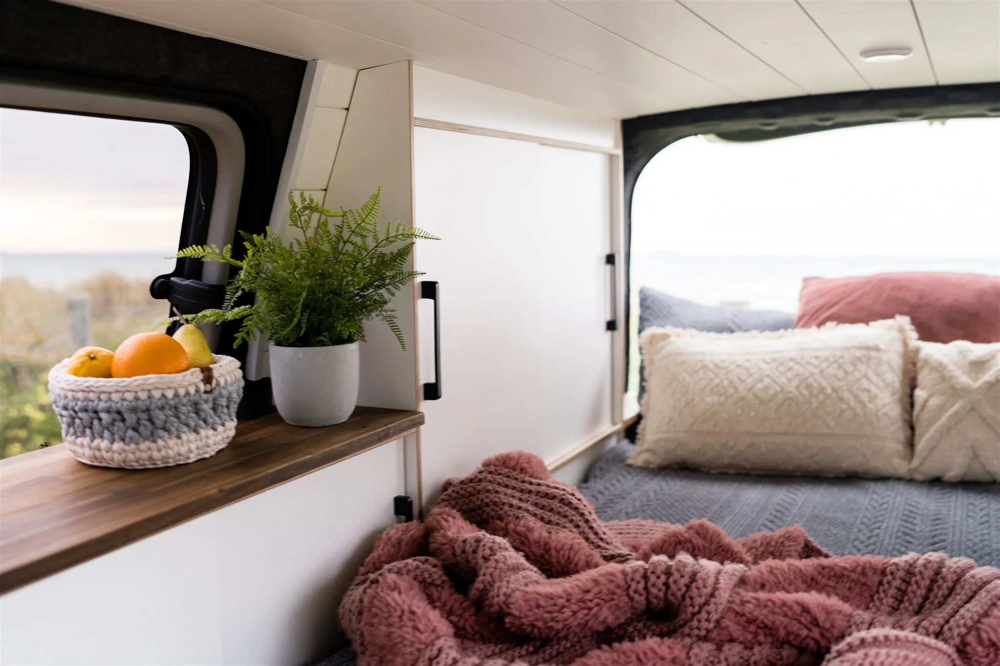 Interior of a camper van with a bed, pillows, and decorative items, with a window showing an outdoor view.