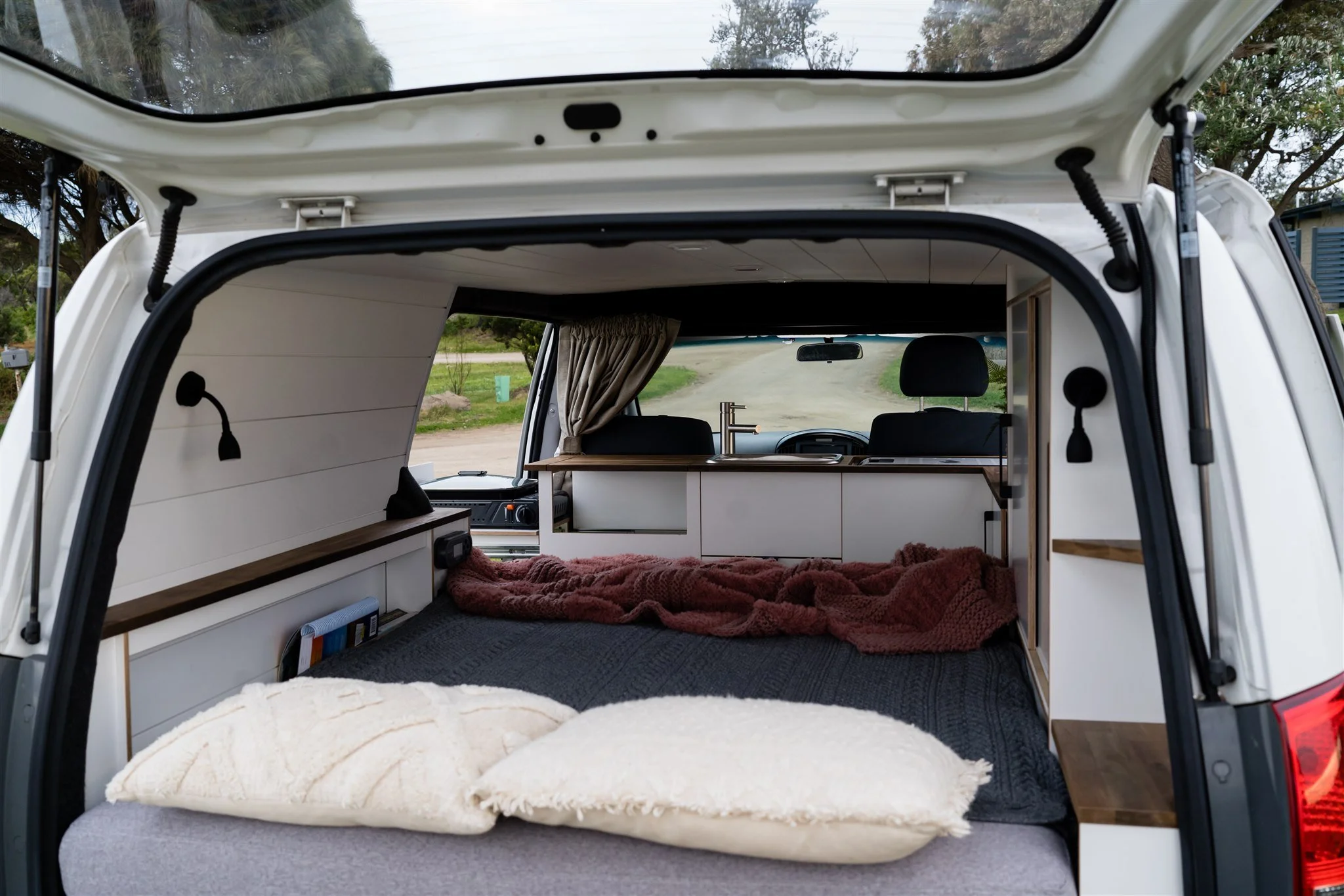 Interior view of a camper van with a bed, pillows, and a small kitchen area, parked on a road with greenery outside.