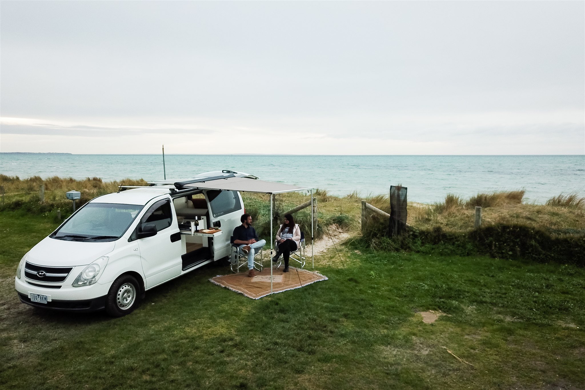 A white camper van parked on grass near the beach with two people sitting outside under an awning, engaging in conversation, with the ocean and cloudy sky in the background.