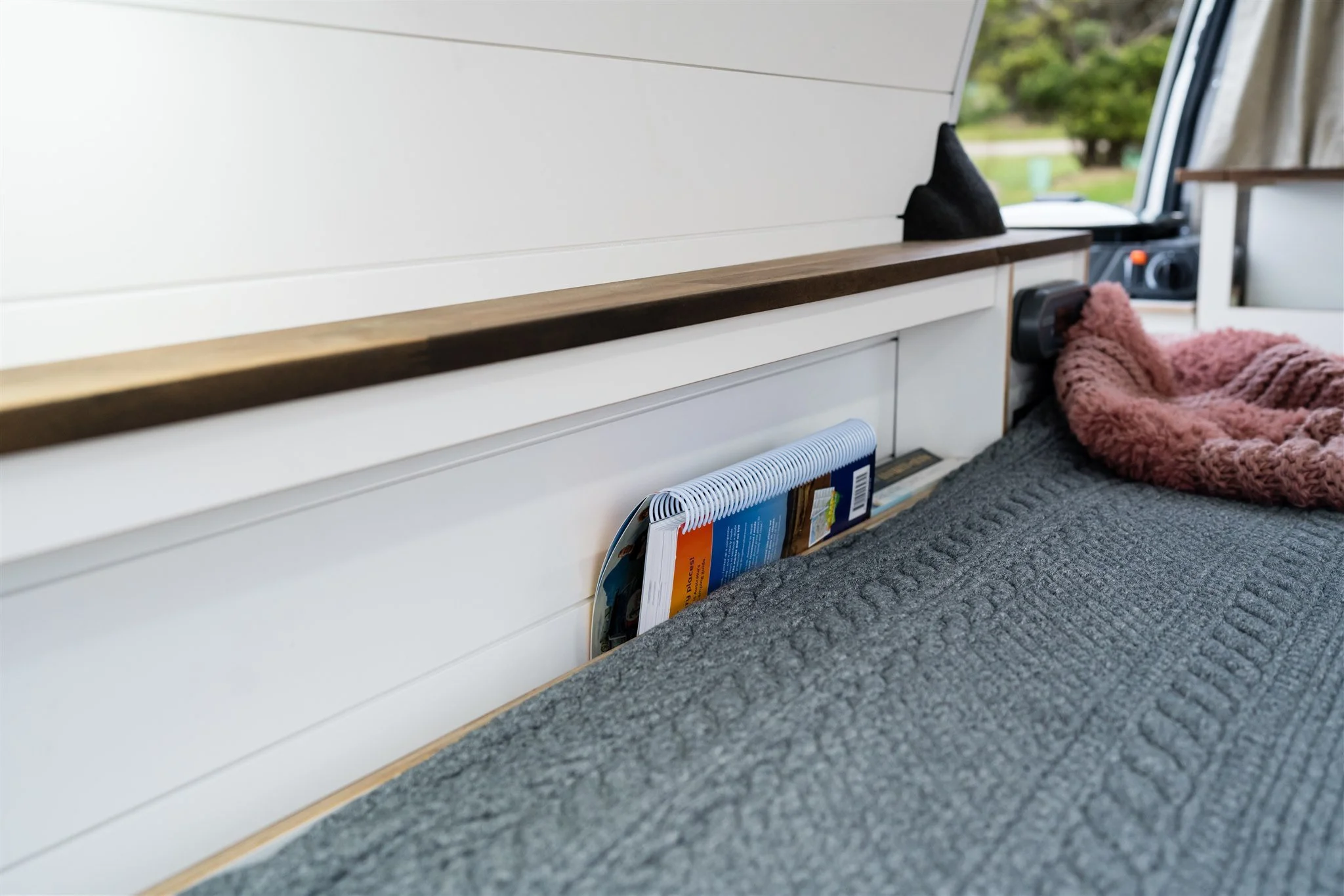 Interior of a camper van with a bed, a book and a spiral notebook stored in a bed side pocket, and a pink blanket on the bed near a window and a control panel.