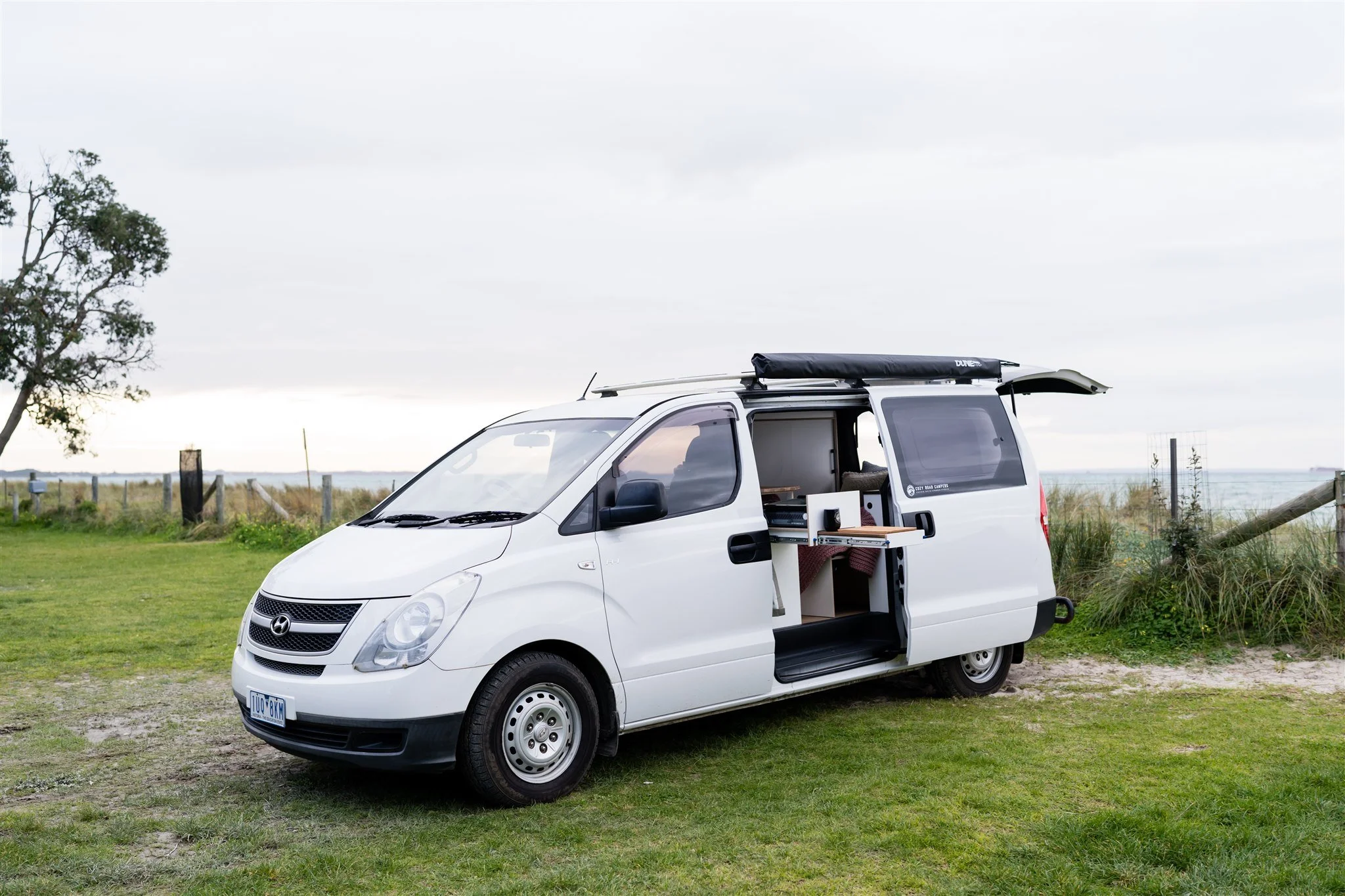 A white camper van parked on grass near a fence with the ocean in the background. The van's side door is open, revealing an interior with a small kitchen and seating area. A surfboard is mounted on the roof.