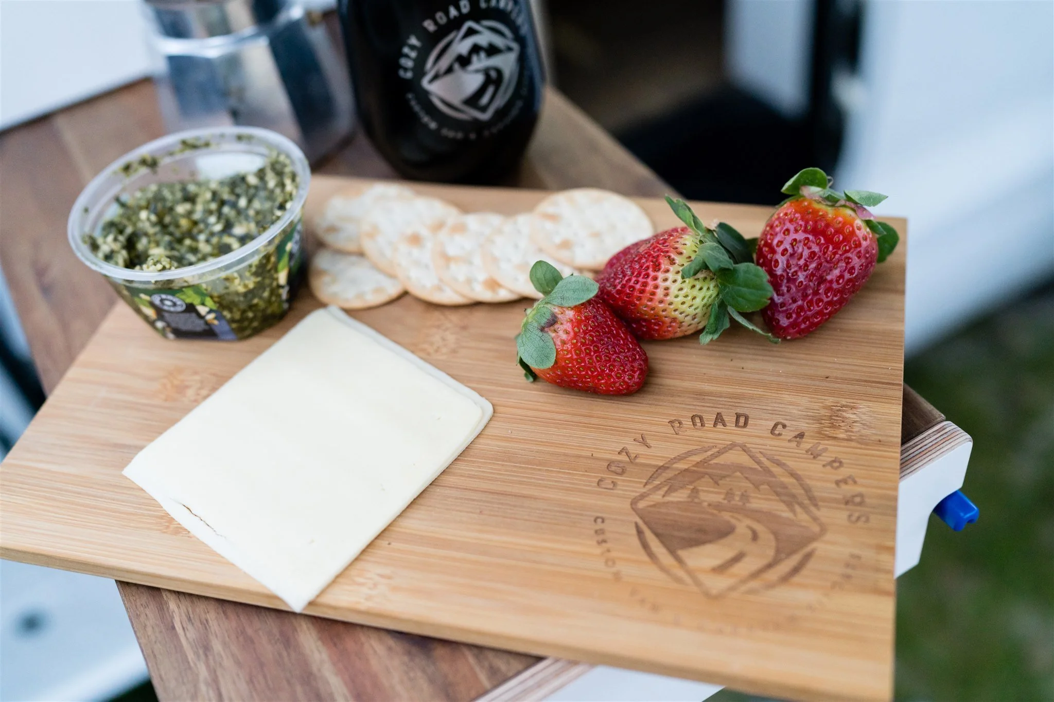 A wooden cutting board with three strawberries, slices of cheese, crackers, a container of pesto, and a bottle of soda.