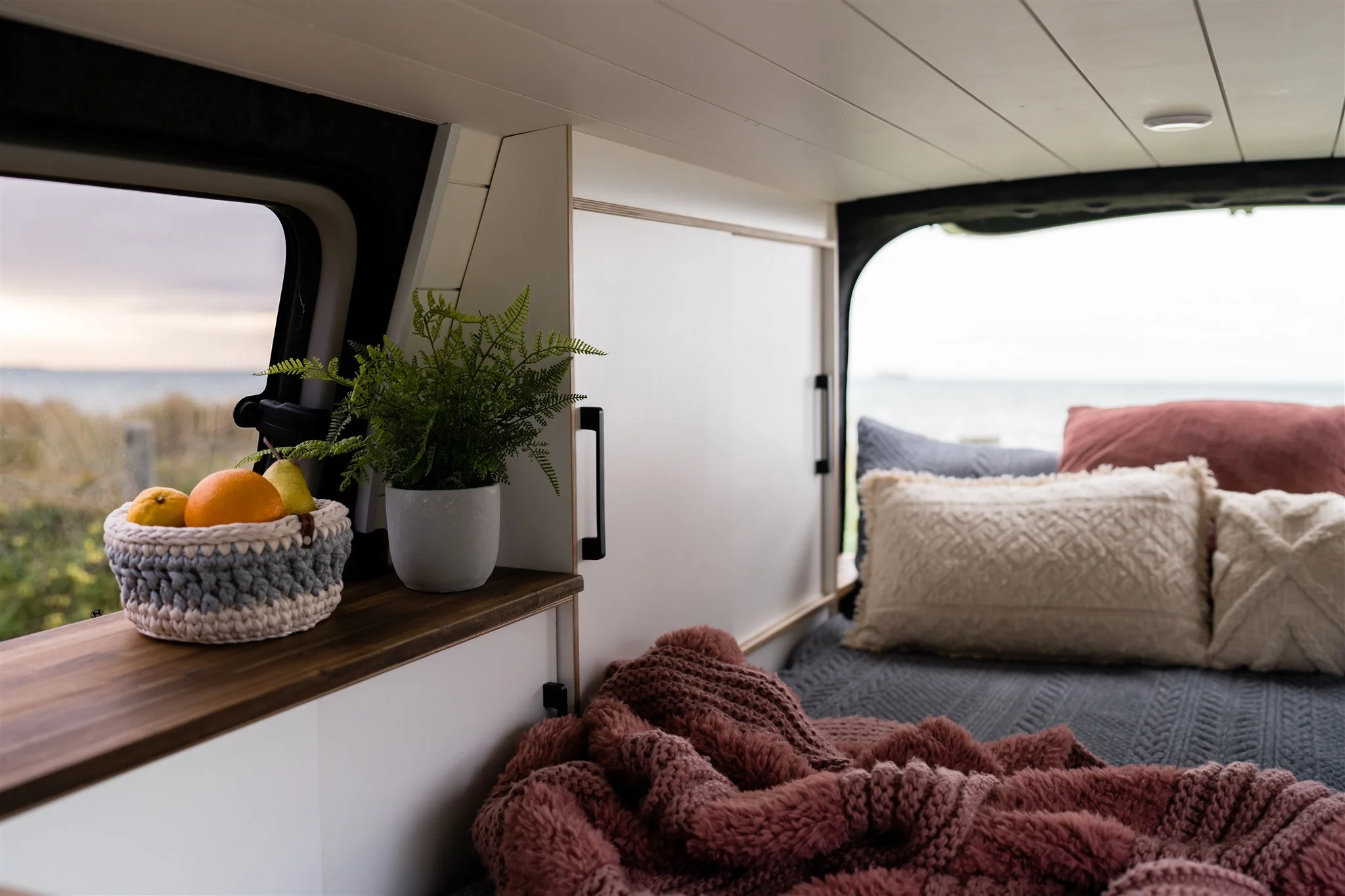Cozy interior of a camper van with a bed, pillows, a window with a scenic view, potted plant, and a basket of fruit.