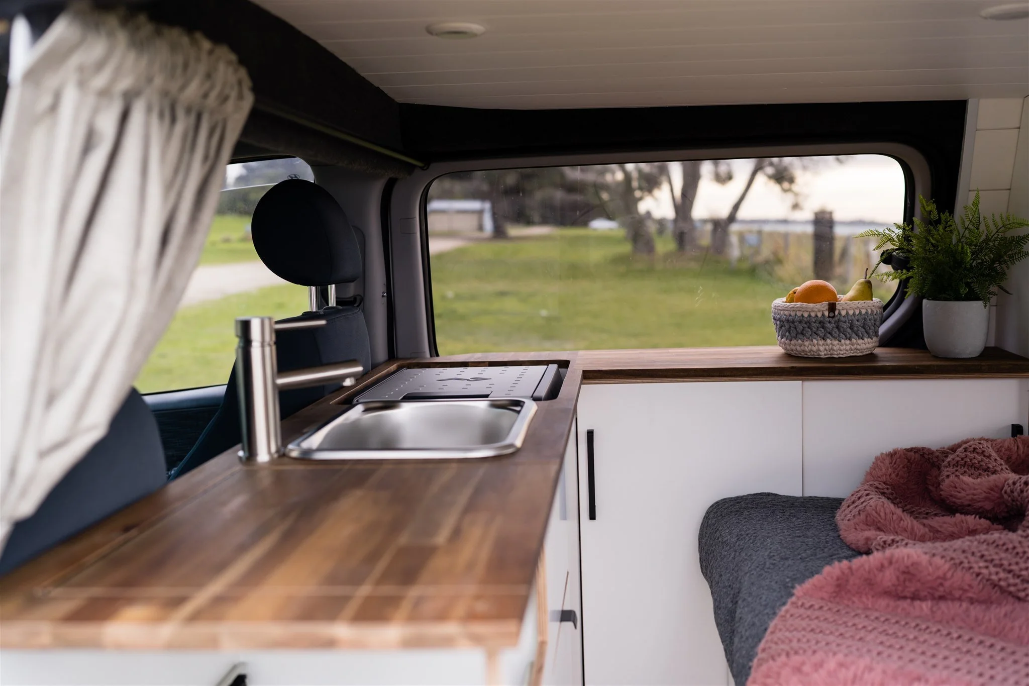 Interior view of a camper van with a wooden countertop, a sink, and a window showing a grassy landscape with trees outside. There are decorative items like a basket of fruit and a potted plant, along with a cozy blanket on a seating area.