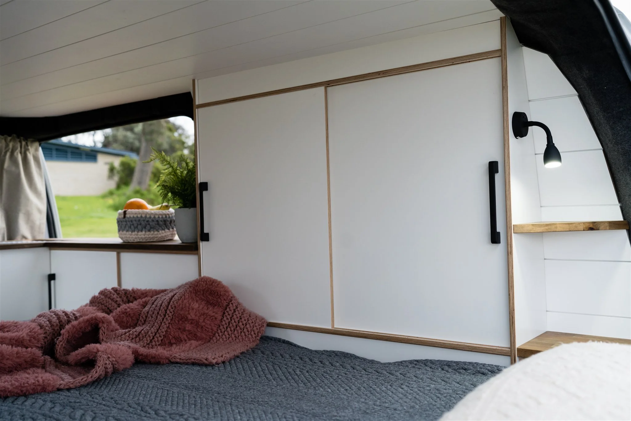 Interior of a cozy camper with white walls, black handles, a window with a curtain, a small shelf, a lamp, a plant, and a bed with a cozy blanket.