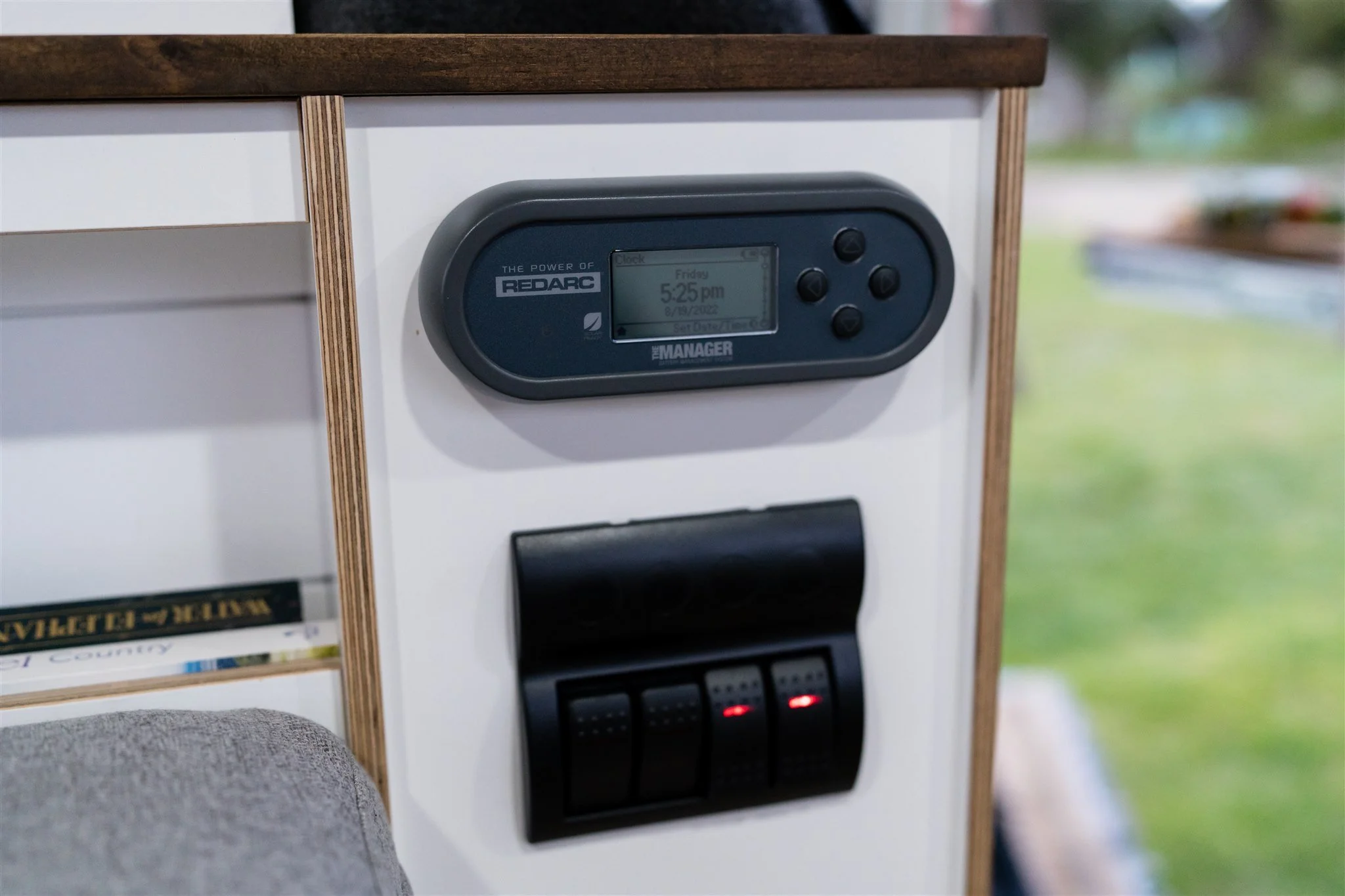 Close-up of a white wall with a digital clock and control panel mounted on it. The digital clock shows the time as 5:25 PM on a Friday, August 18, 2002. The control panel below has four black switches with red indicator lights.