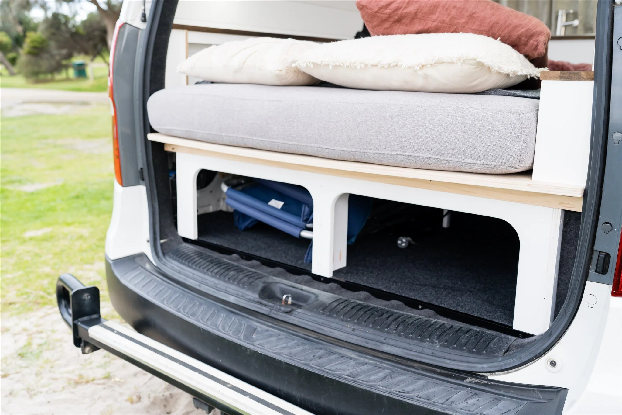 The back of a white van with a built-in bed and storage area, showing cushions, pillows, and rolled-up camping gear inside.