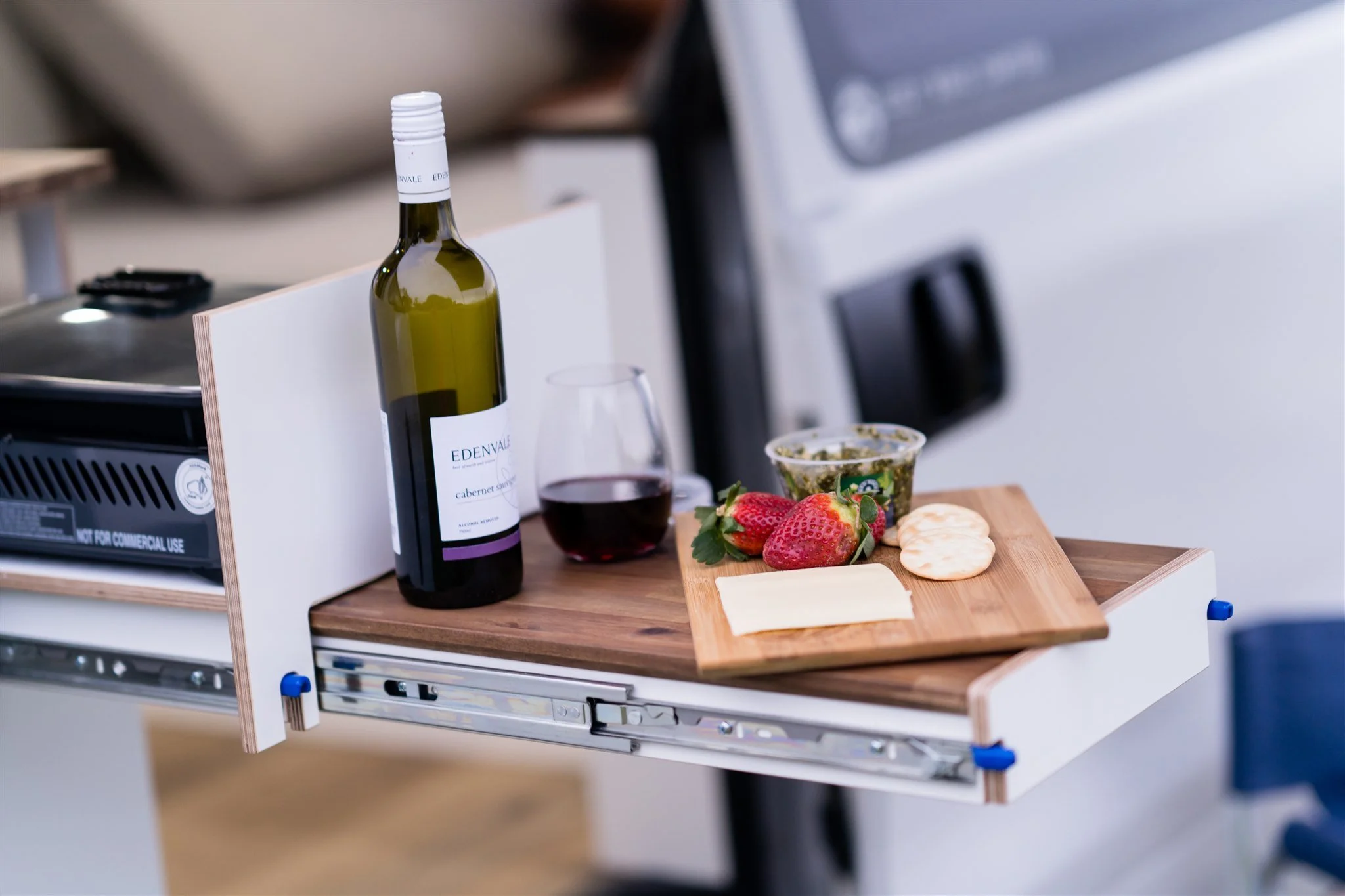 A pull-out tray with a bottle of red wine, a glass of red wine, strawberries, cheese, crackers, and a small container of green spread.