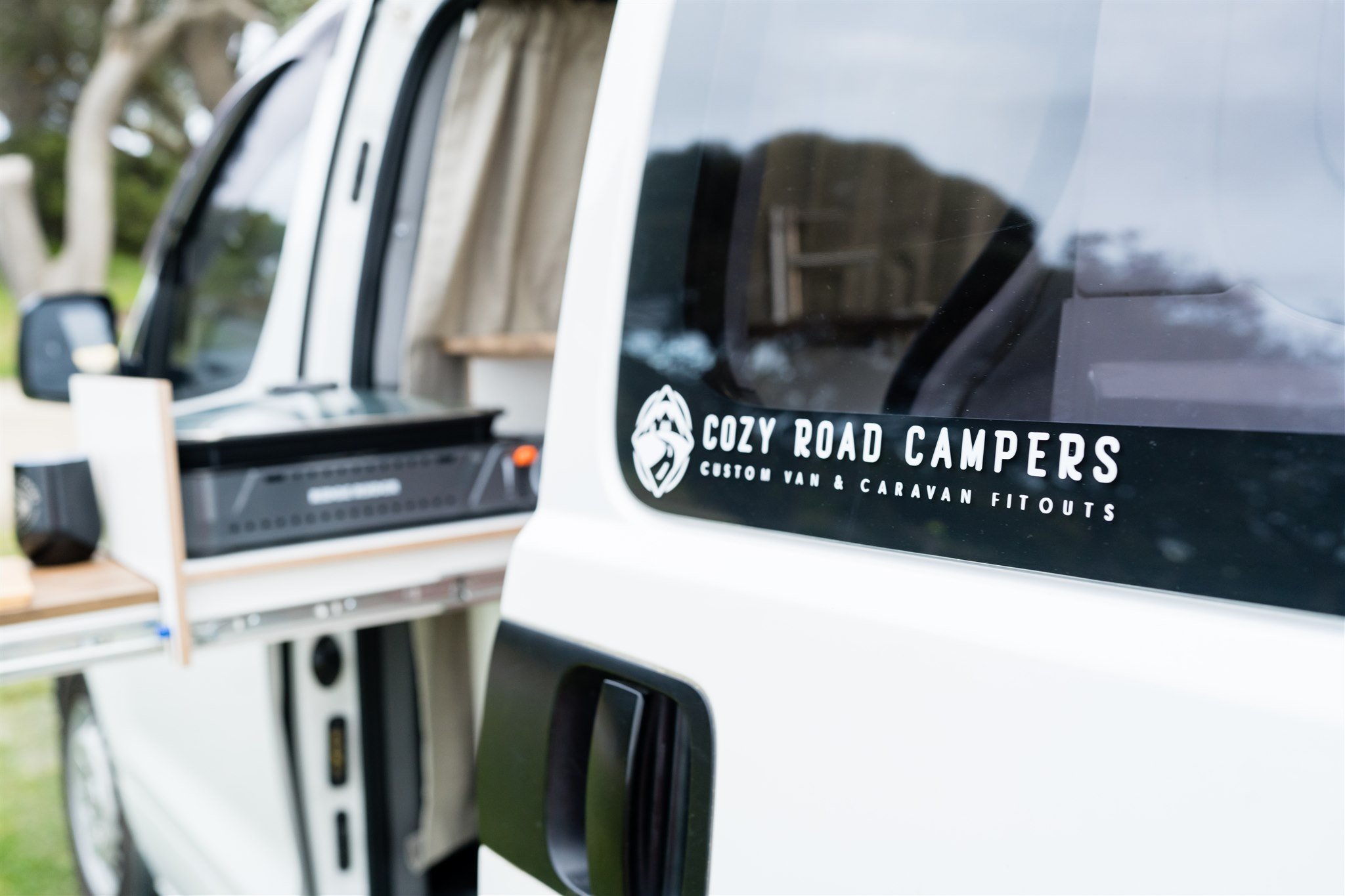 Close-up of a white camper van with a decal that reads 'Cozy Road Campers, Custom Van & Caravan Fitouts' on the rear window, showing a blurred outdoor shed and greenery in the background.