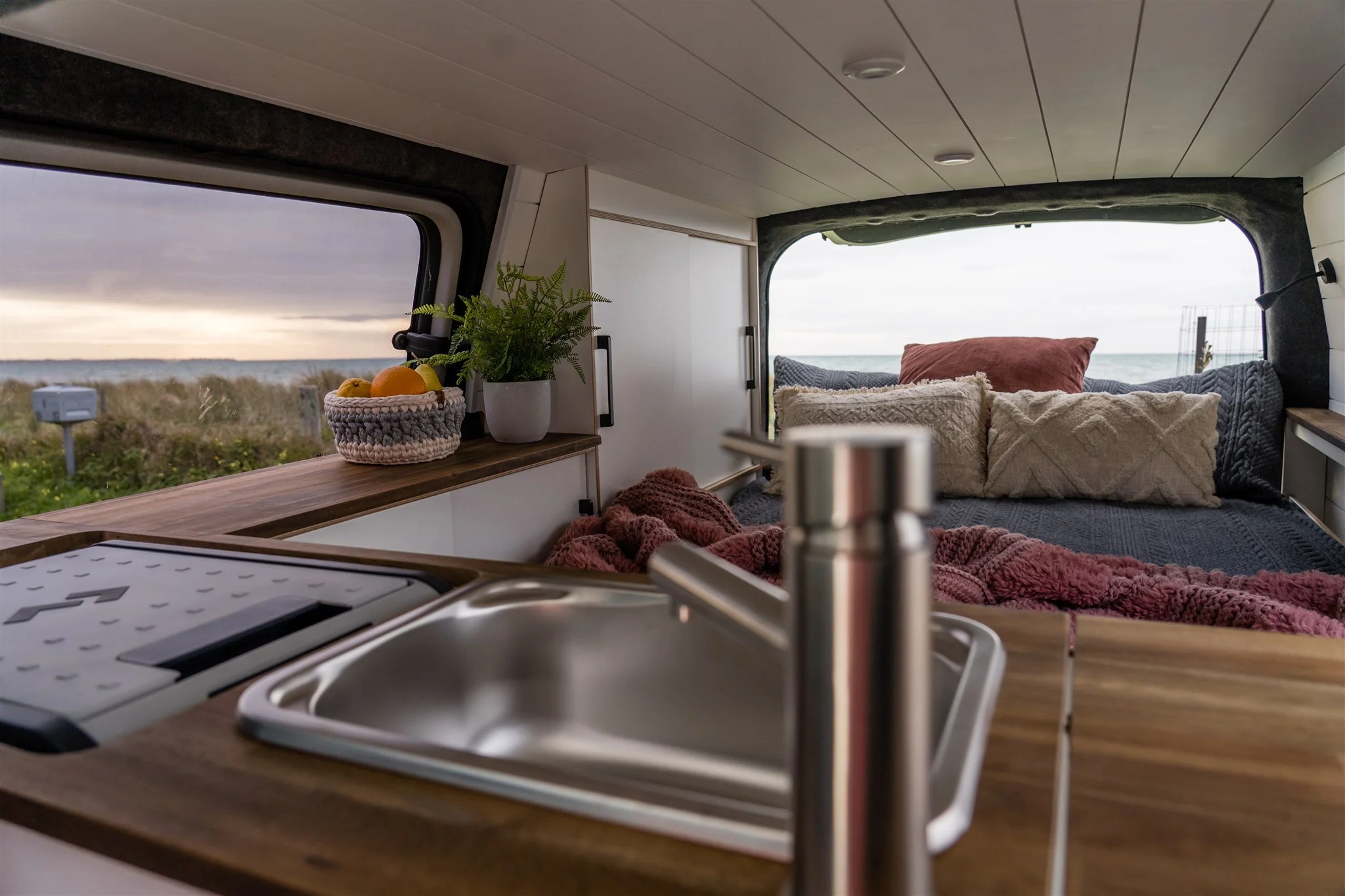 Interior of a camper van with a view of the beach through the windows; kitchen sink and countertop in foreground, decorated with a plant and a basket of fruit, and a cozy bed with pillows in the background.