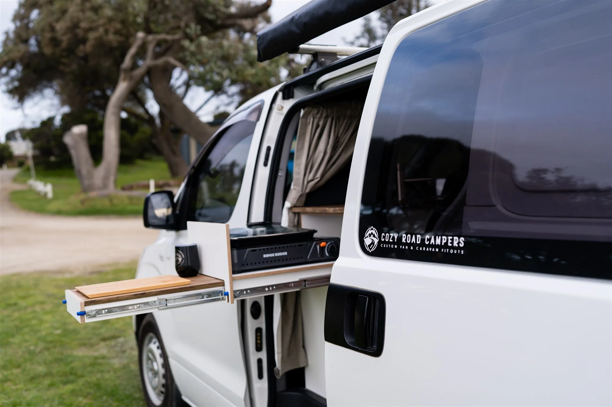 White camper van with an extendable wooden tray attached to the side, a small kitchen area inside with curtains, set outdoors on grass near trees.