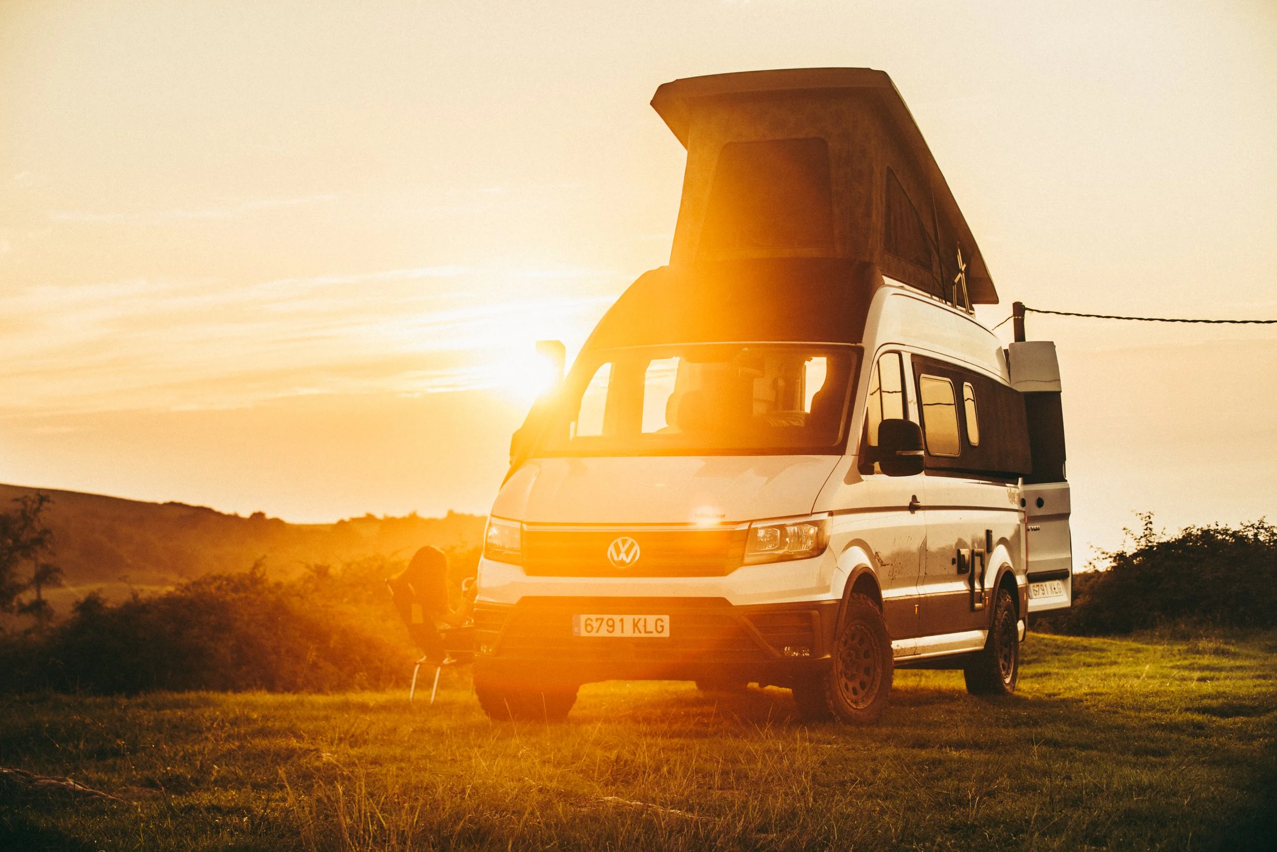 A white camper van with a pop-up roof parked on a grassy field during sunset, with a person sitting on a chair nearby.