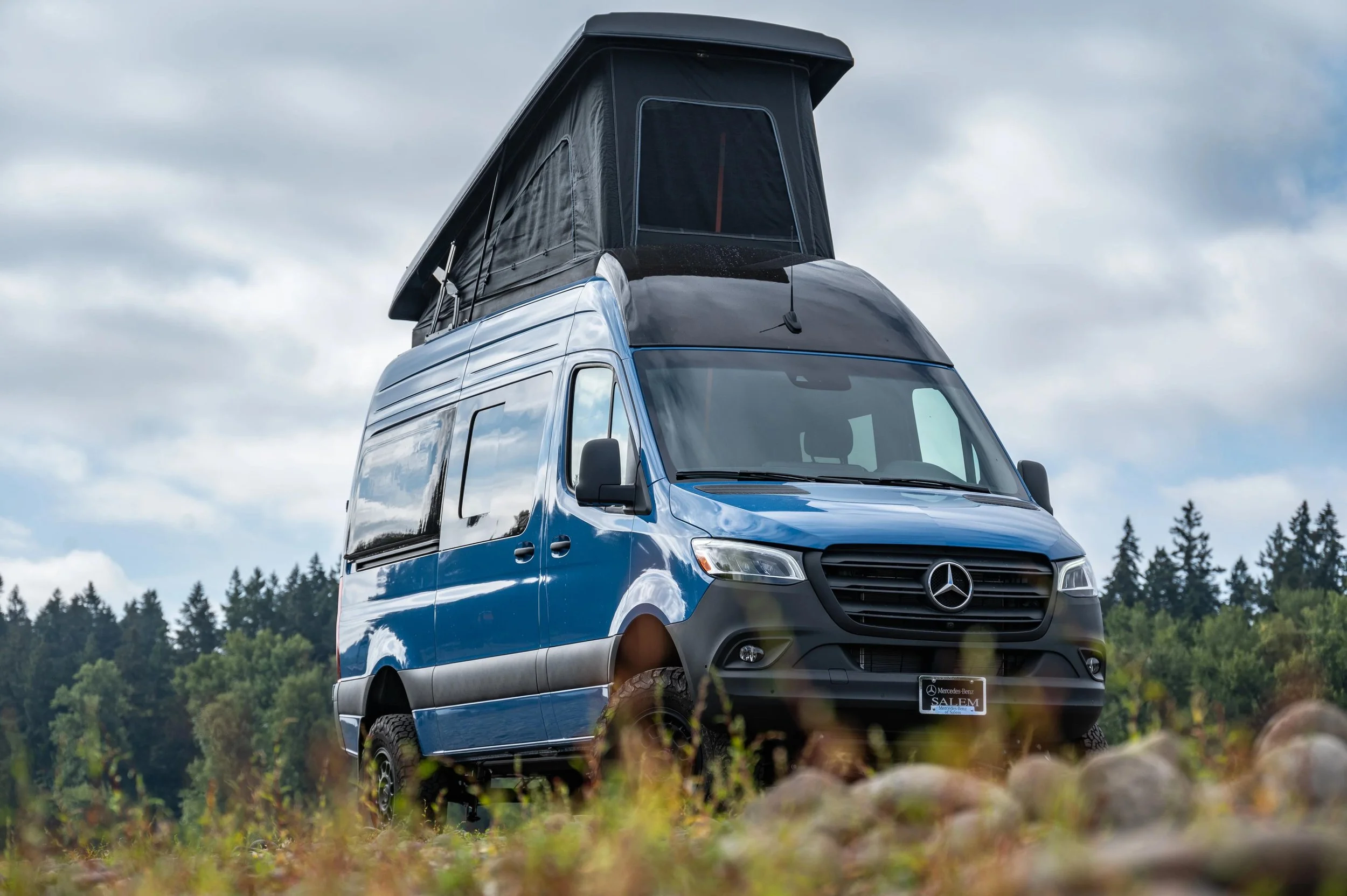 Blue Mercedes-Benz camper van with a pop-up roof, parked outdoors on a rocky and grassy area with forest and cloudy sky in the background.