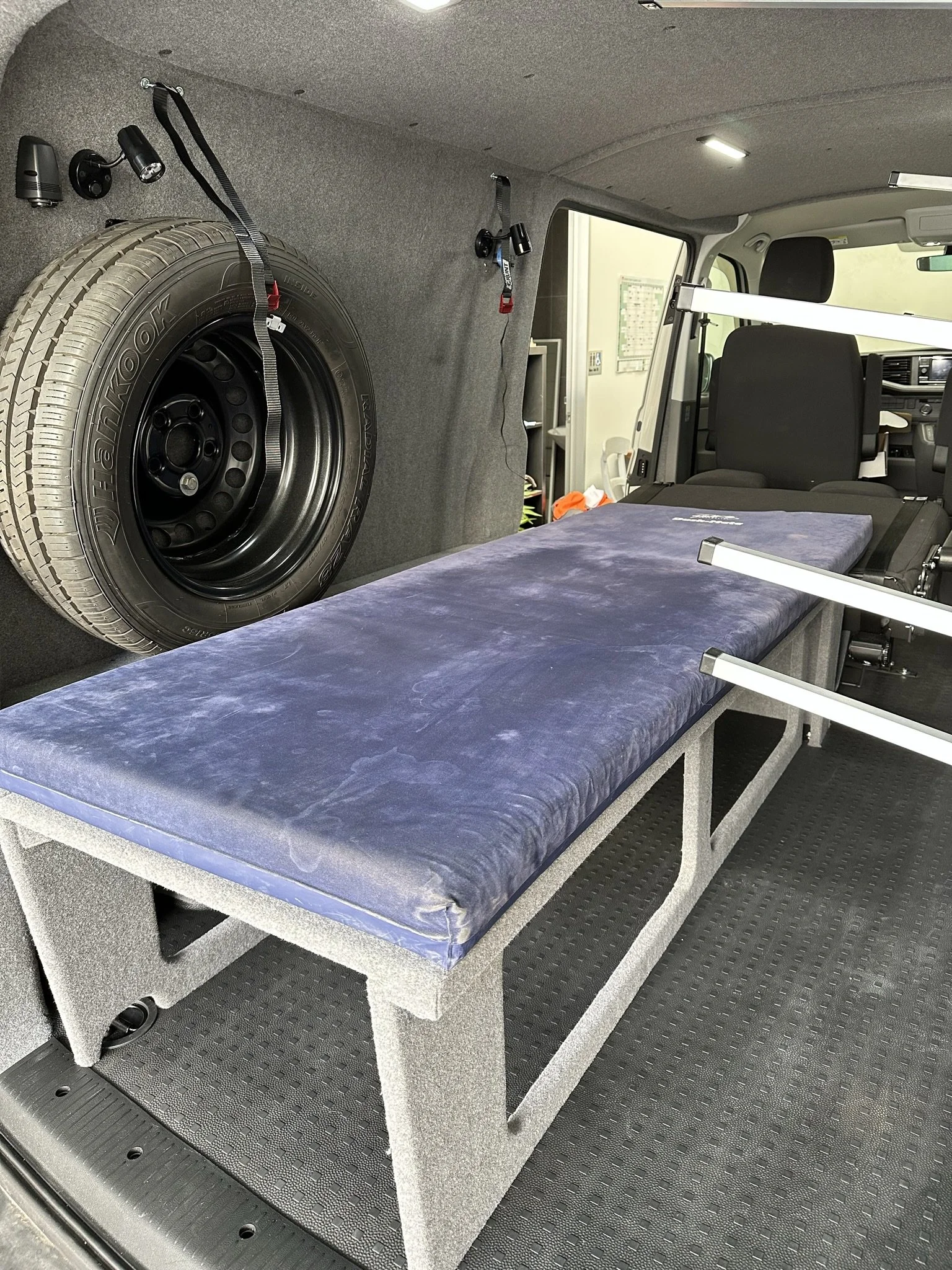 Interior of a van converted for mobility or medical use, featuring a bench with a cushioned surface, a mounted spare tire, and straps on the wall for securing equipment.