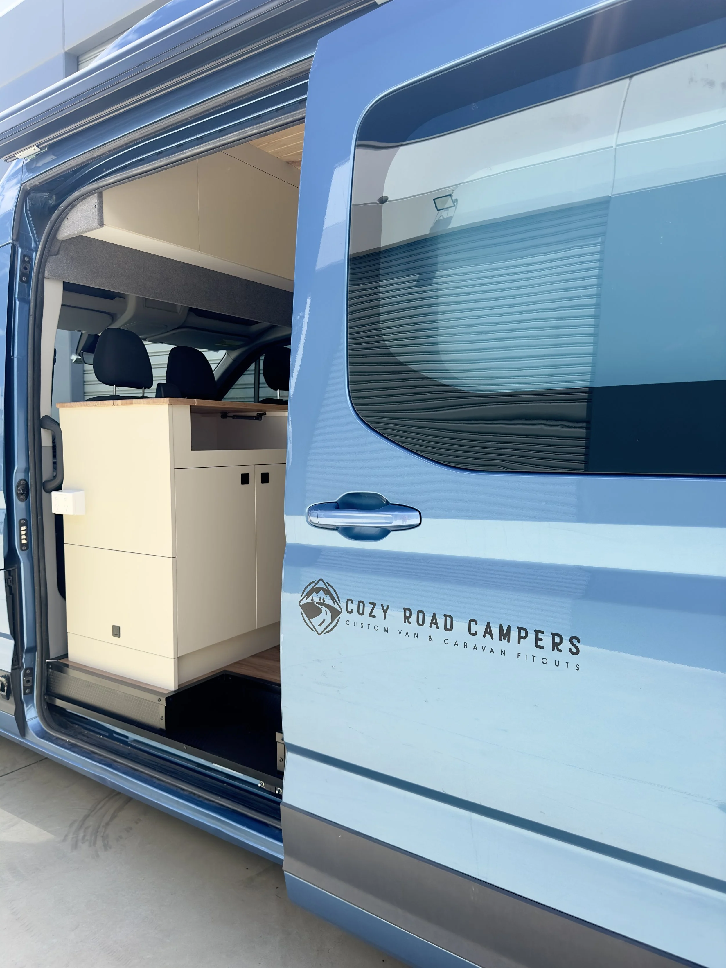 Open sliding door of a blue camper van labeled Cozy Road Campers with a partial view of interior cabinets and seats.
