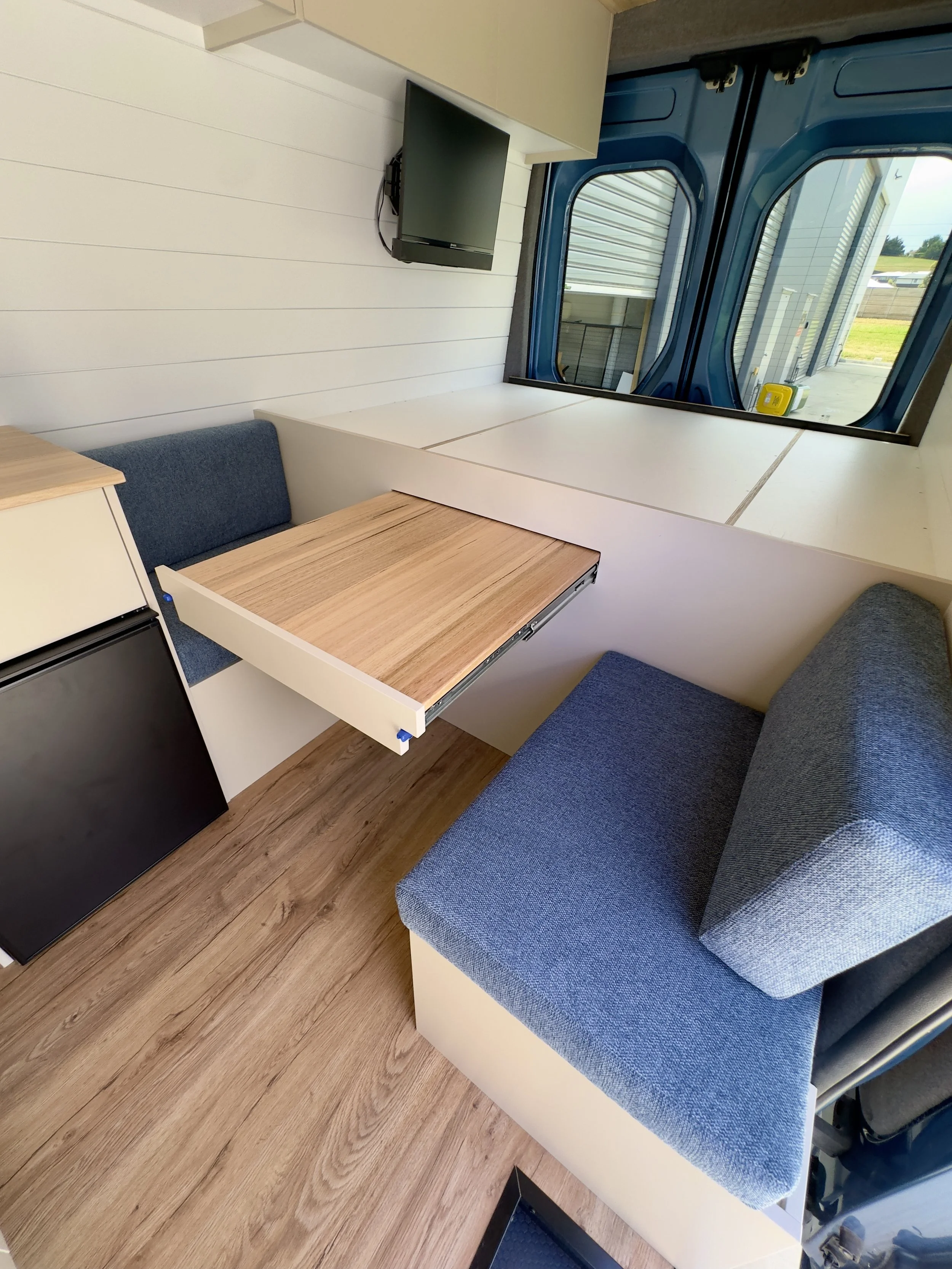 Interior of a small vehicle or cabin with a wall-mounted television, a fold-out wooden table, a blue cushioned seat, windows showing an outdoor scene, and a small refrigerator.