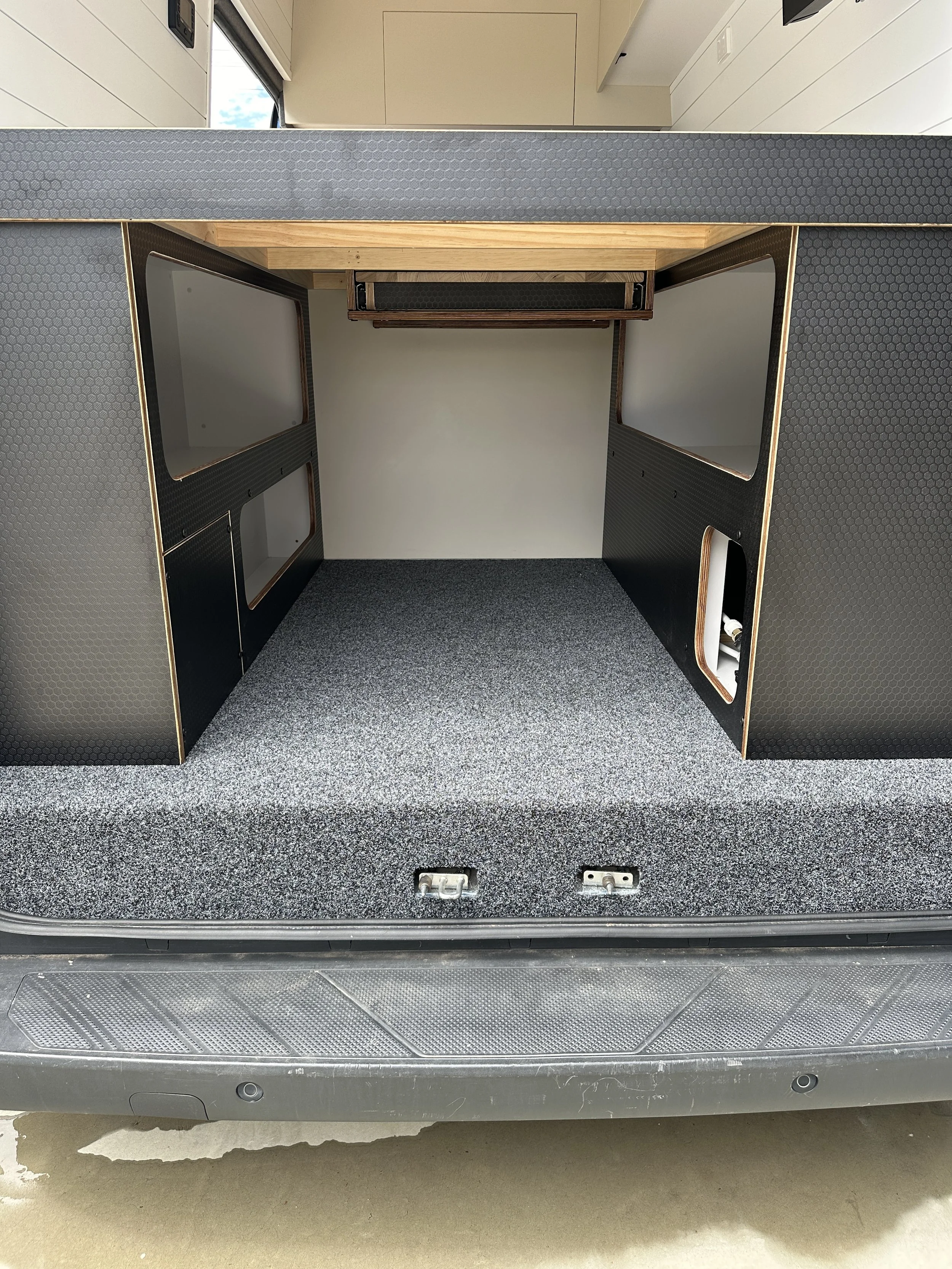 Empty custom-built van conversion with black textured side panels, various cutouts, a wooden work surface, and a carpeted floor area, viewed from the rear.