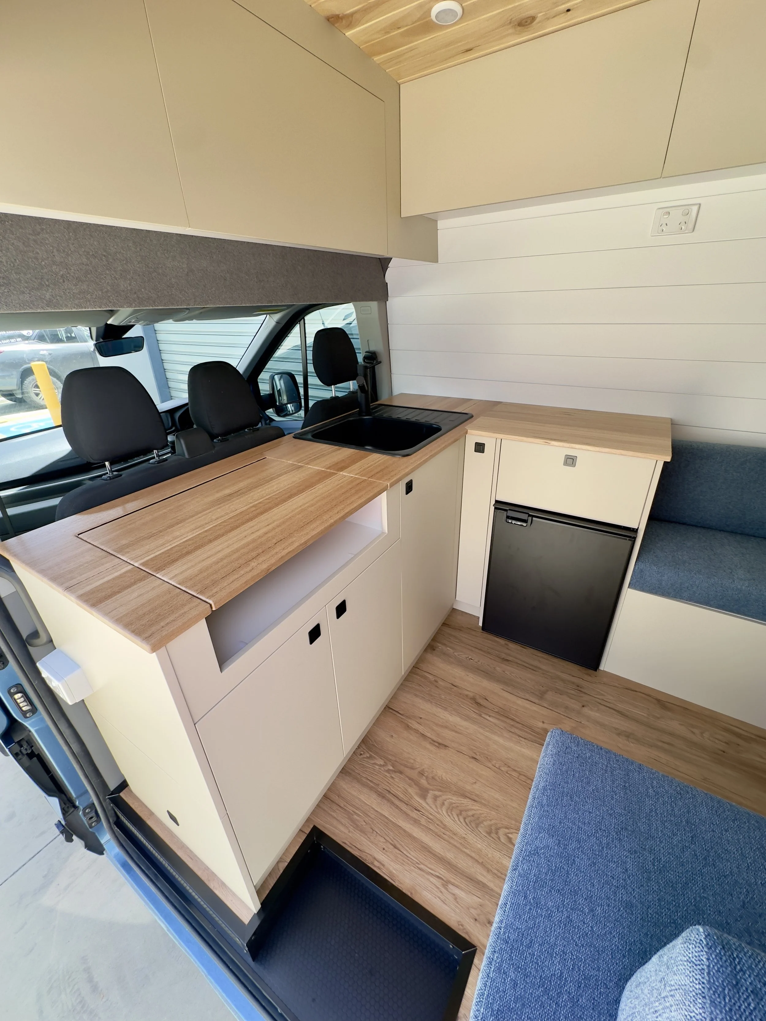Interior of a compact camper van kitchen with a small black refrigerator, a wooden countertop, a black sink, overhead storage cabinets, and a white wall with a power outlet.