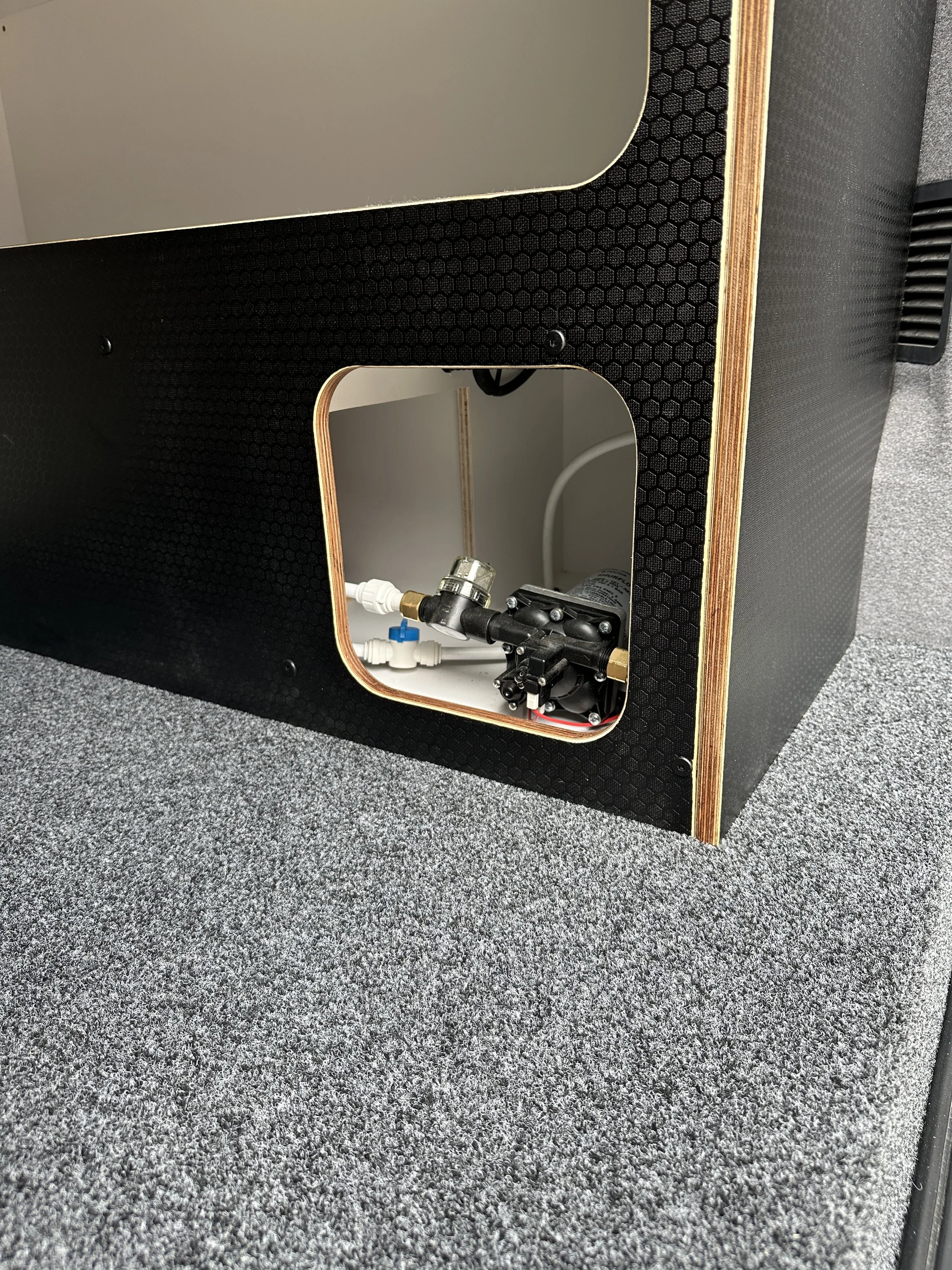 Close-up of the back of a speaker showing an opening with internal components, including pipes and a small motor, on a carpeted floor.