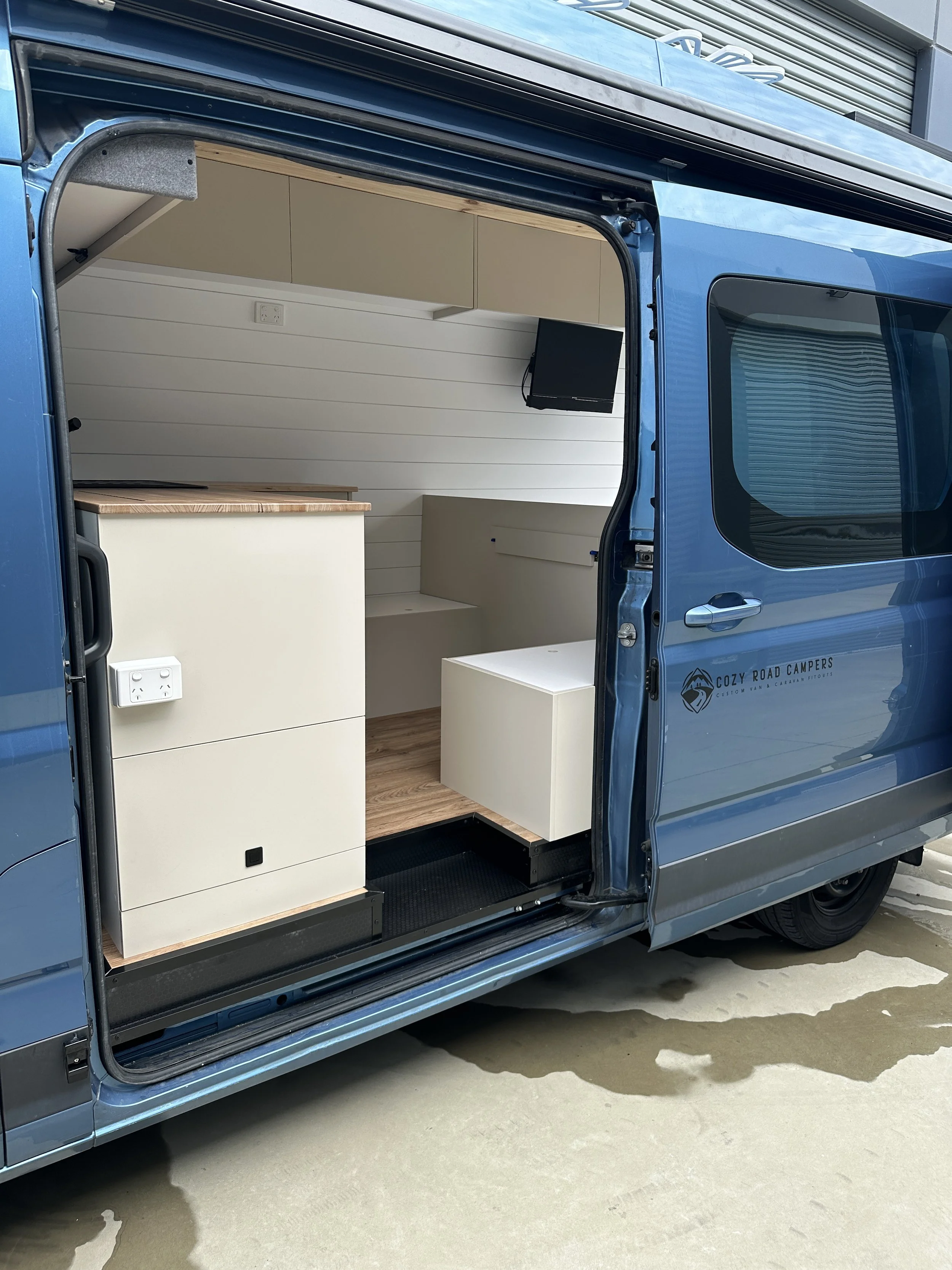 Interior of a blue camper van, showing a small kitchen area with white cabinets, a small TV mounted on the wall, and a beige and wood-colored bench seat.