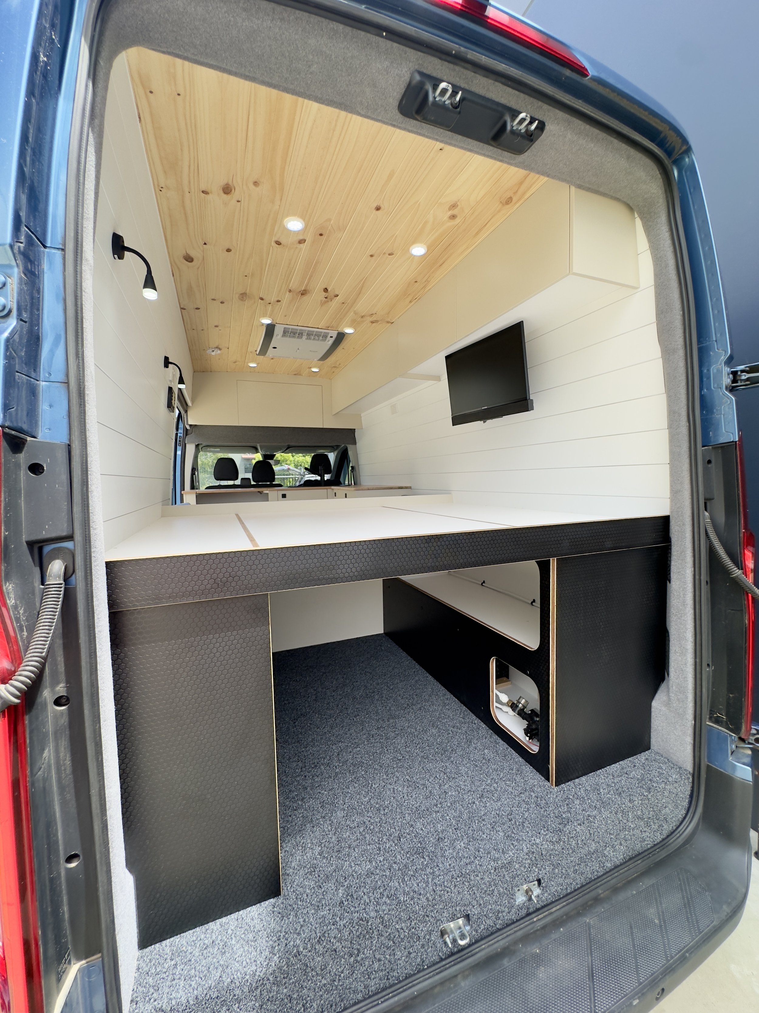 Interior of a converted van with wooden ceiling, white walls, a mounted TV, and a black and white built-in desk underneath the TV.