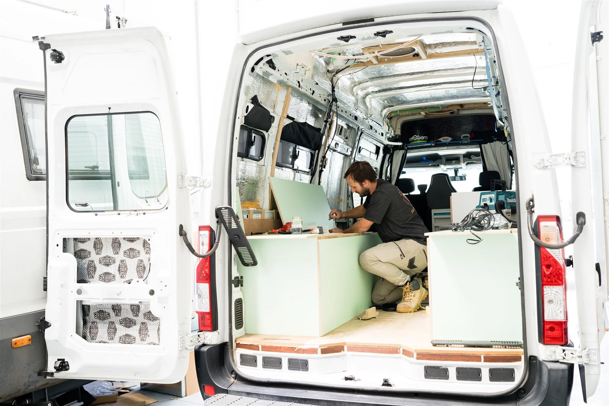 A man working on a green interior panel inside a white van that is being converted into a camper or mobile workspace.