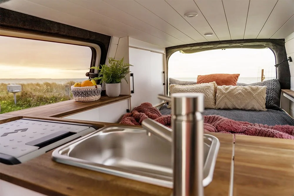 Interior of a campervan with a view of a bed area with pillows, a small sink, and a window showing an outdoor landscape.