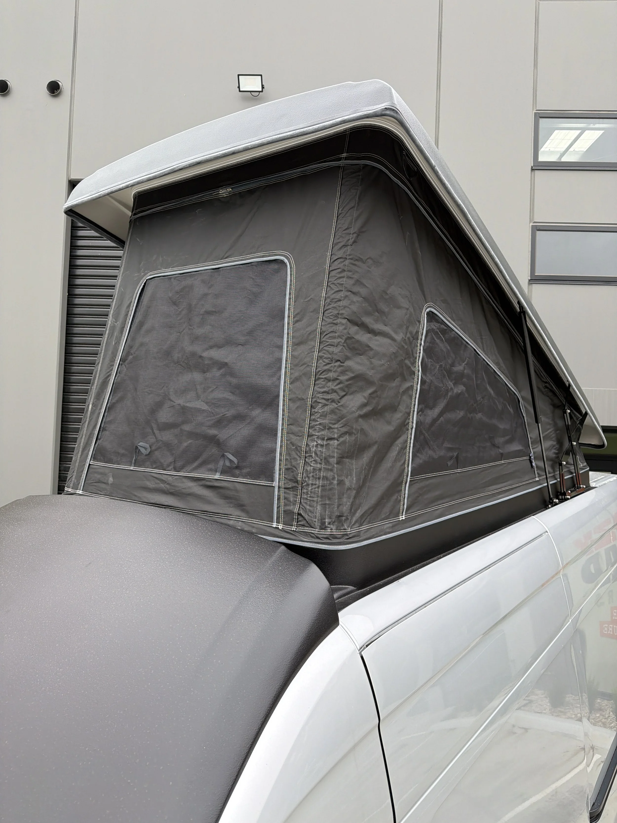 A campervan with a gray rooftop bed, parked in front of a building with gray walls and windows.