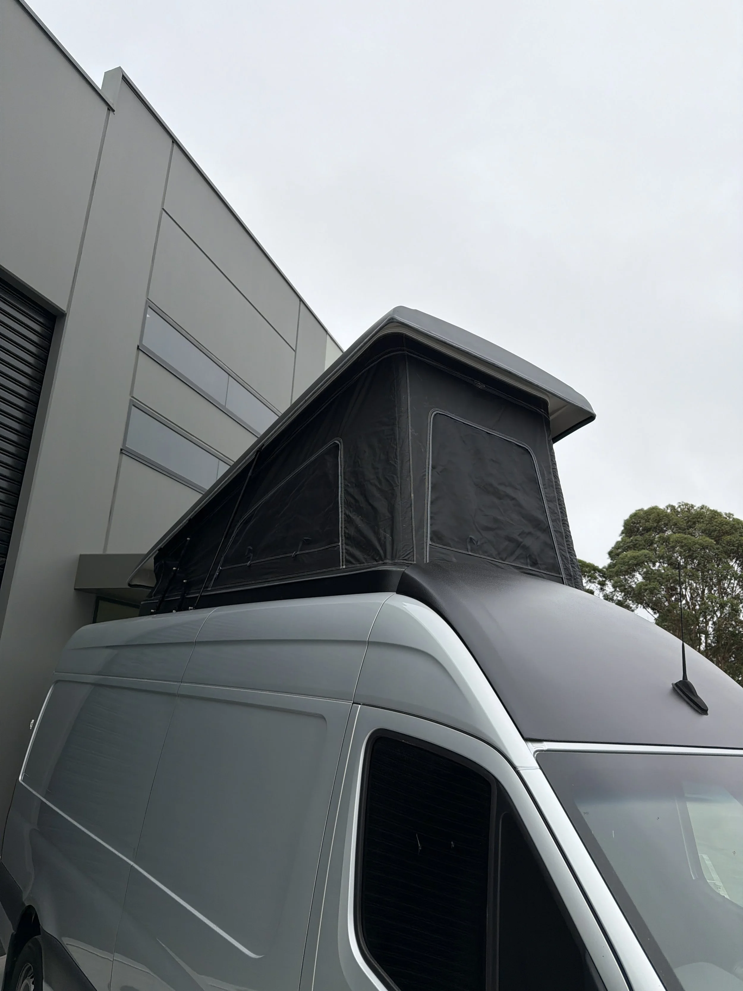 A white van with a black tent or canopy on top, parked beside a modern building with grey panels and windows, under an overcast sky, with trees visible in the background.