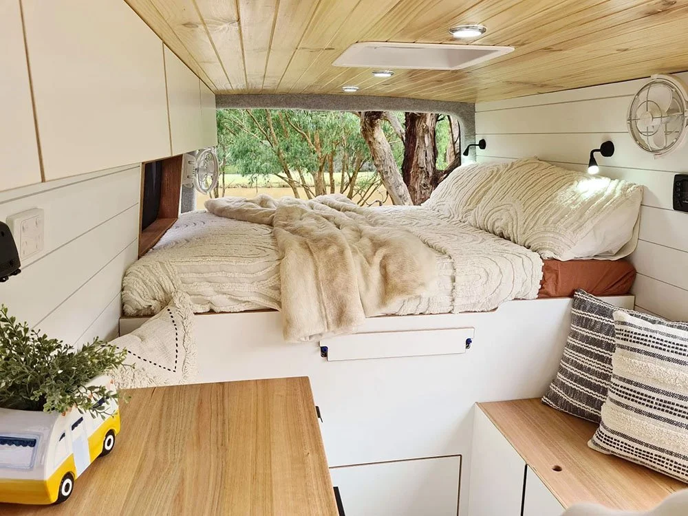 A cozy bedroom with a bed inside a small camper or van, overlooking trees outside through an open window. The bed has cream-colored bedding, a faux fur blanket, and pillows. There's a wooden surface with a potted plant and a small yellow and white camper toy. The interior features wood paneling and built-in lighting.
