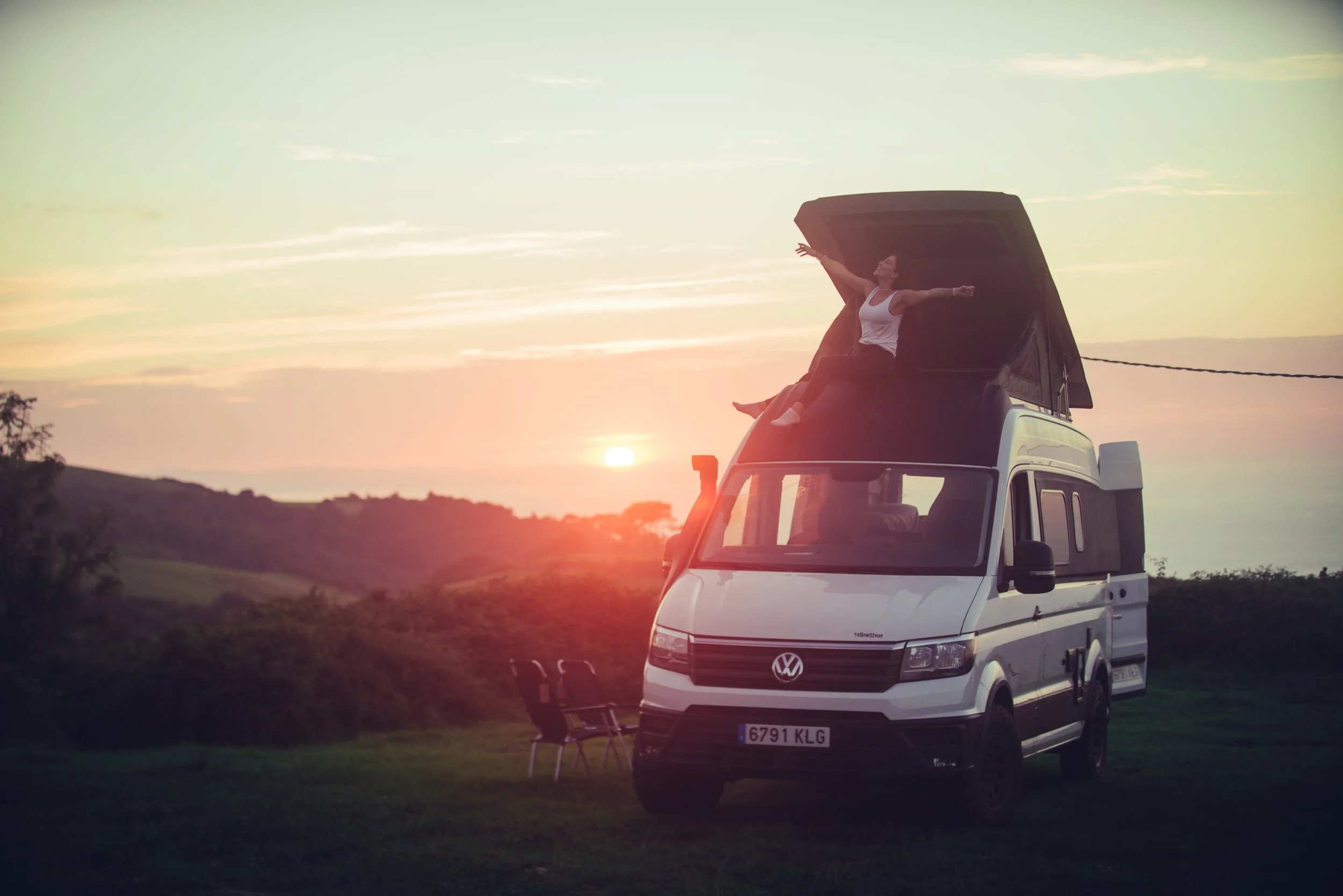 A woman sitting on the edge of an open van's roof, with arms outstretched, during a sunset in a rural landscape.