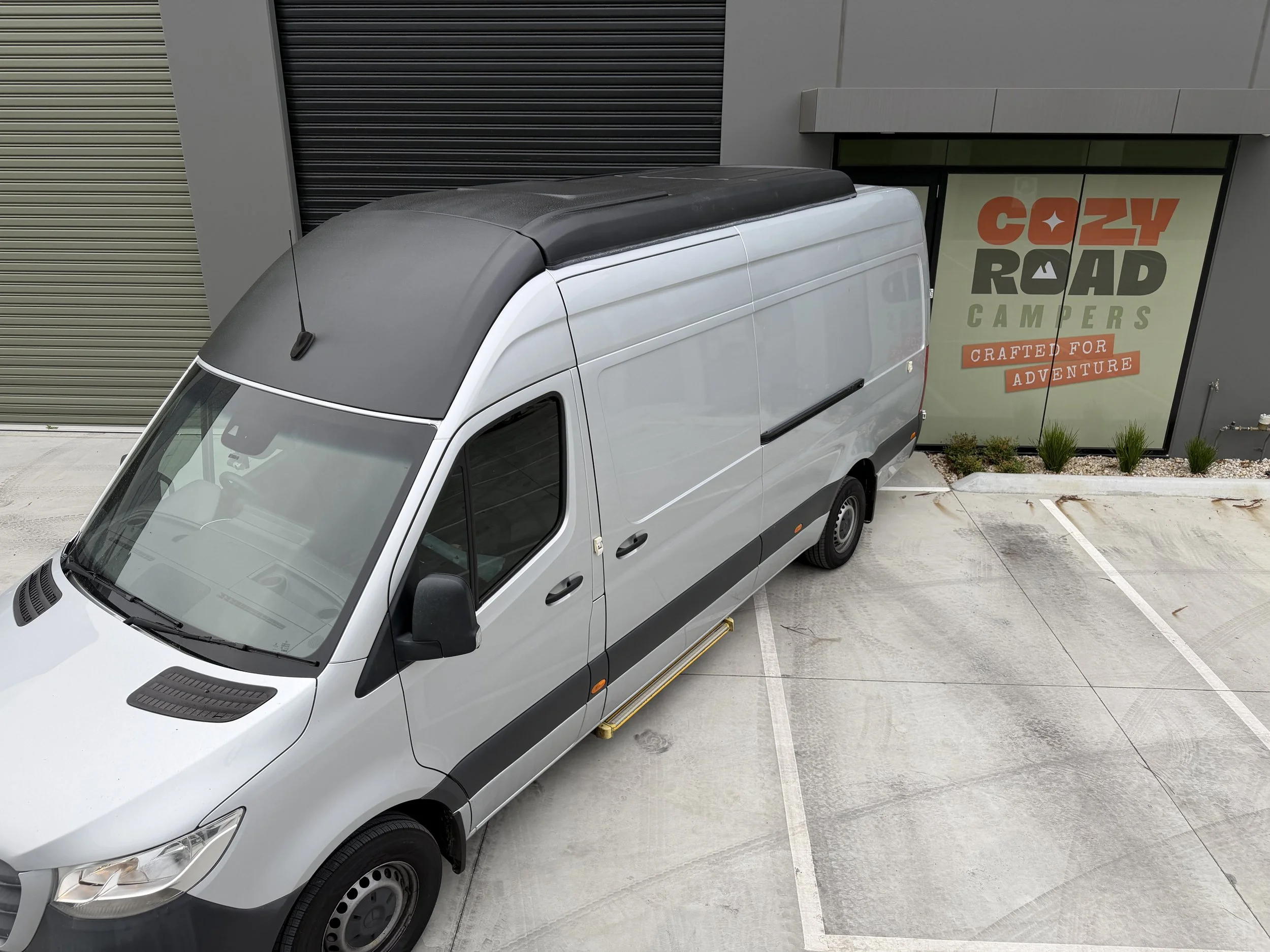 A silver cargo van parked in an empty parking lot outside a building with signage that reads 'Cozzy Road Campers Crafted for Adventure.'