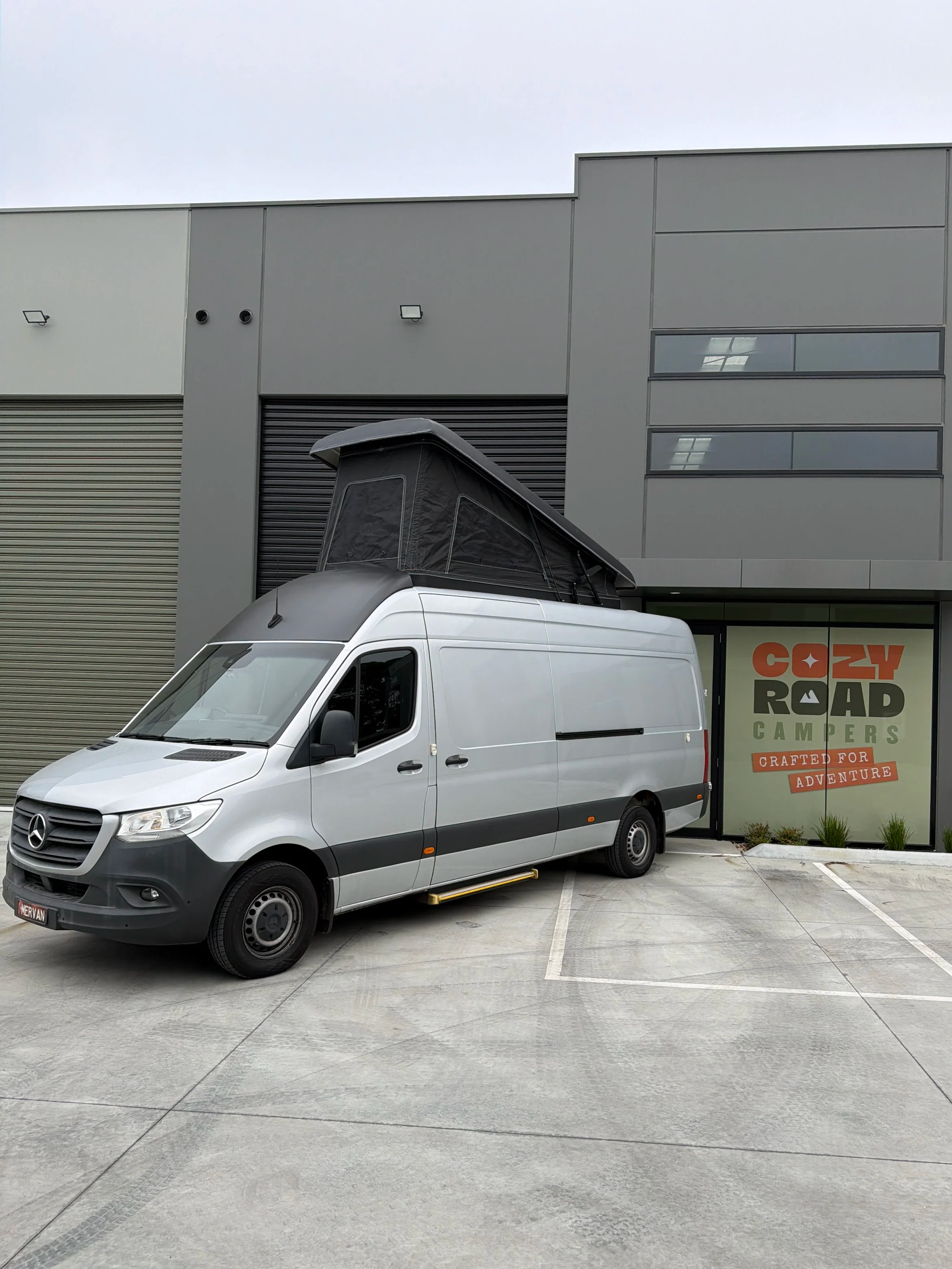 A white Mercedes-Benz Sprinter van with a rooftop tent parked in front of a modern commercial building. The building has gray panels, glass windows, and a sign for Cozy Road Campers that reads "Crafted for Adventure."