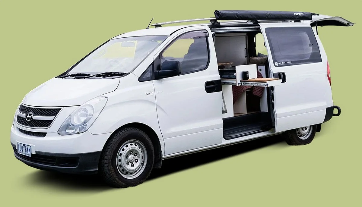 White camper van with the side door open showing a small interior with a foldable table and seating, against a solid light green background.